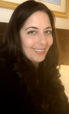 Smiling woman with long dark hair in a black top, sitting indoors near framed wall art.