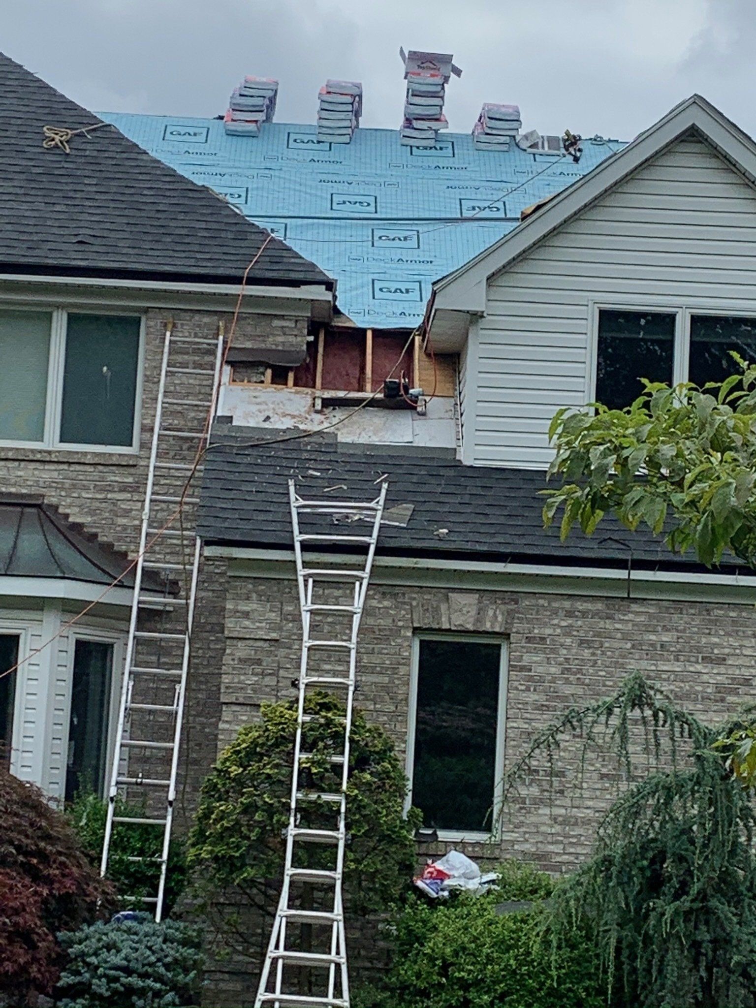 Two-story house with roof under repair, blue underlayment exposed and ladders leaning against the front.