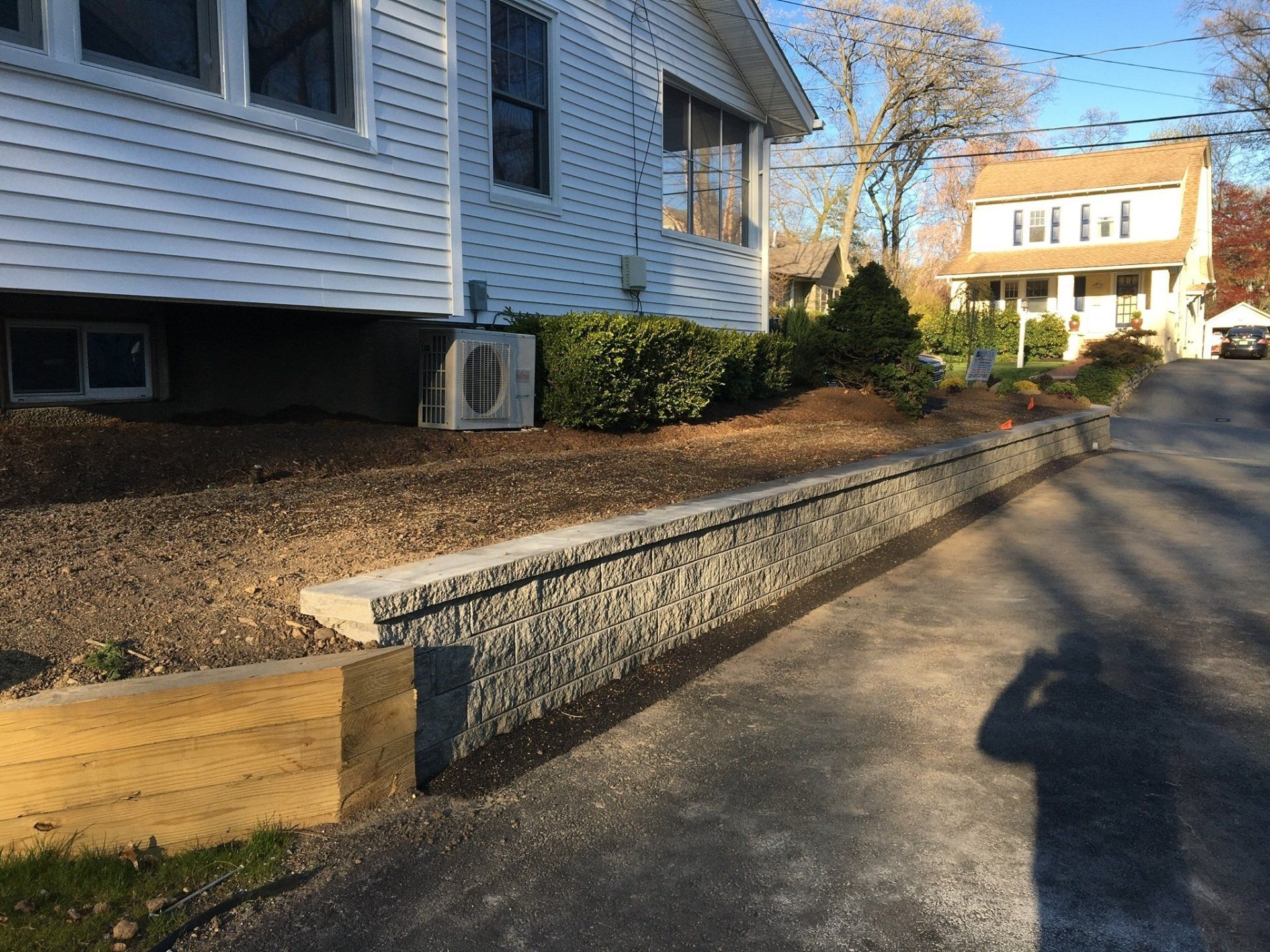 Residential street with houses, a raised stone retaining wall, and a sunlit sidewalk shadow