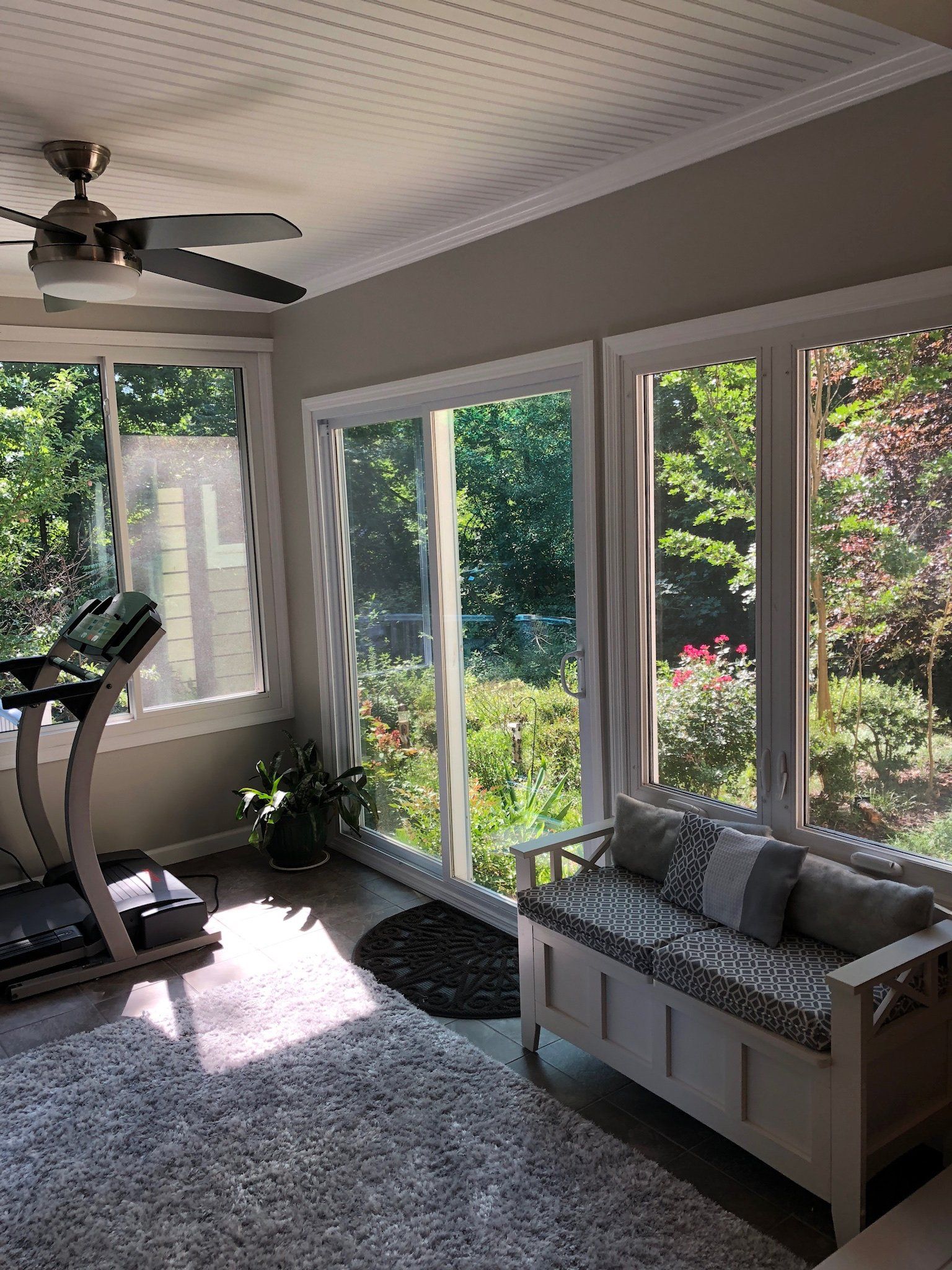Sunlit home gym with treadmill, ceiling fan, and bench beside large windows overlooking greenery