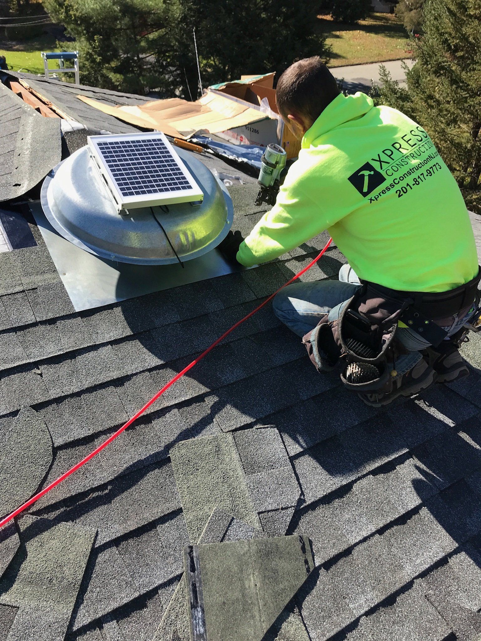 Worker inspecting a roof vent on a shingled rooftop, wearing a neon green safety shirt.