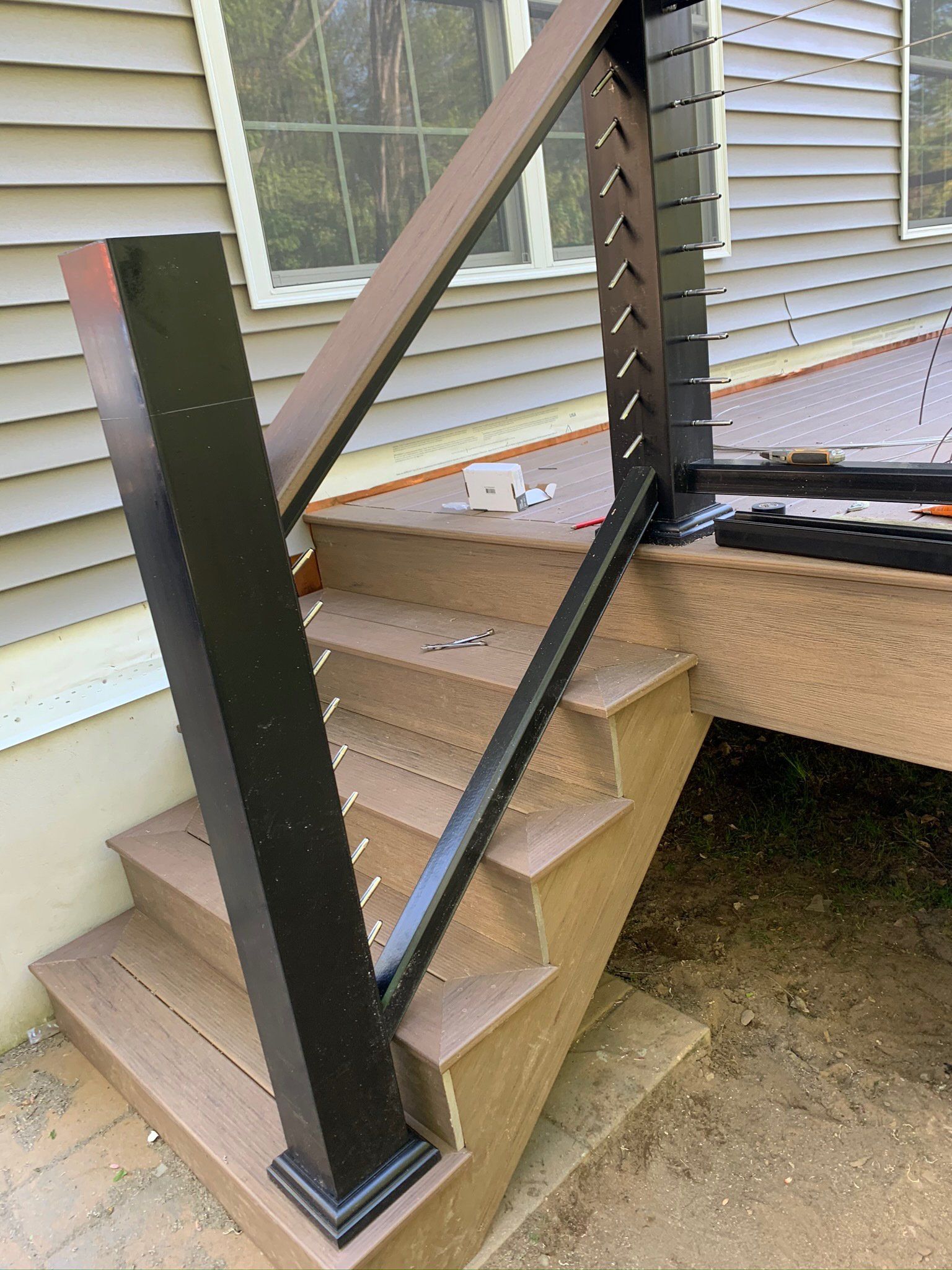 Exterior stairs with black railings and tan steps beside a house siding and deck.