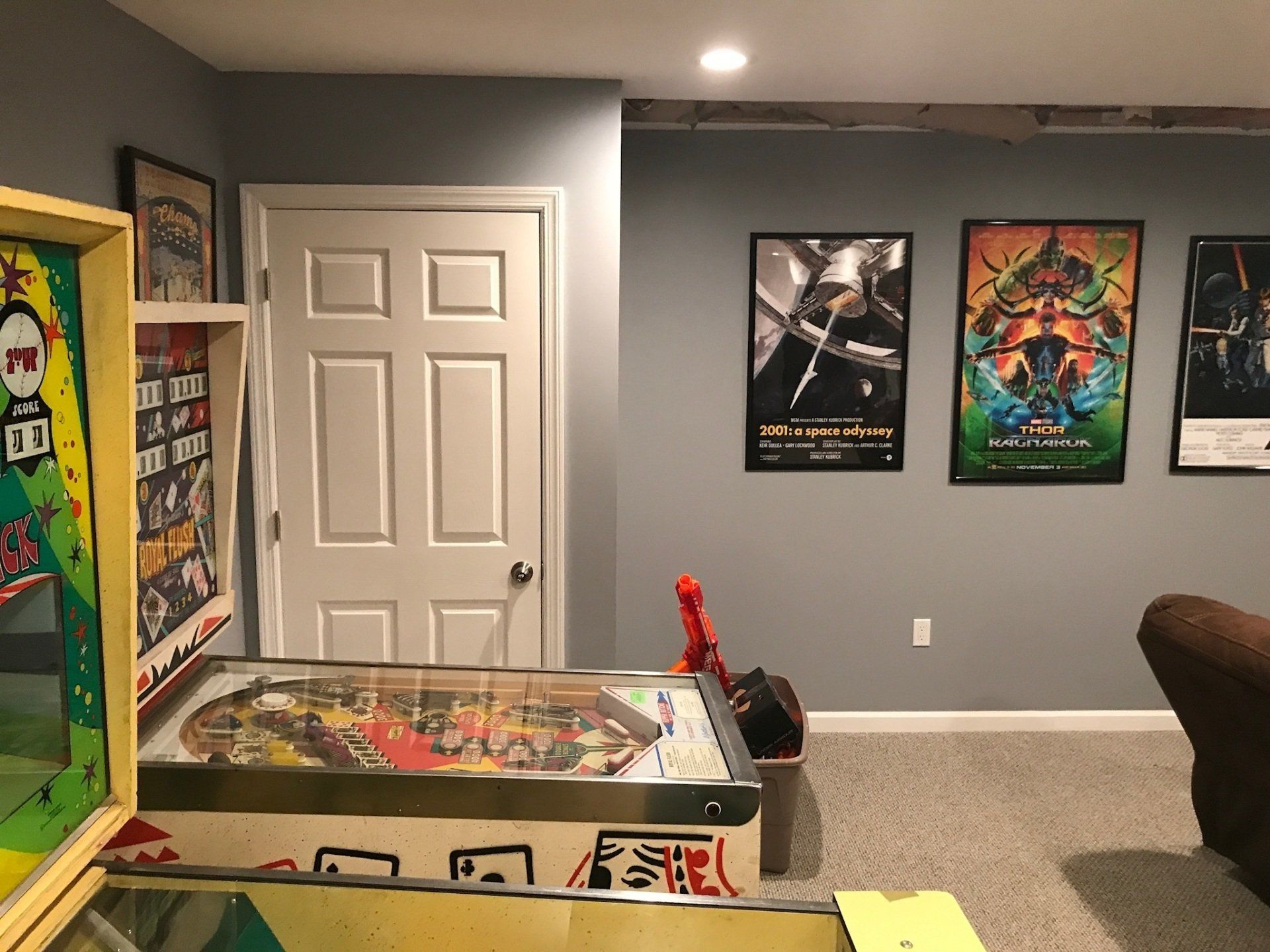 Basement game room with pinball machine, white door, and three movie posters on a gray wall.