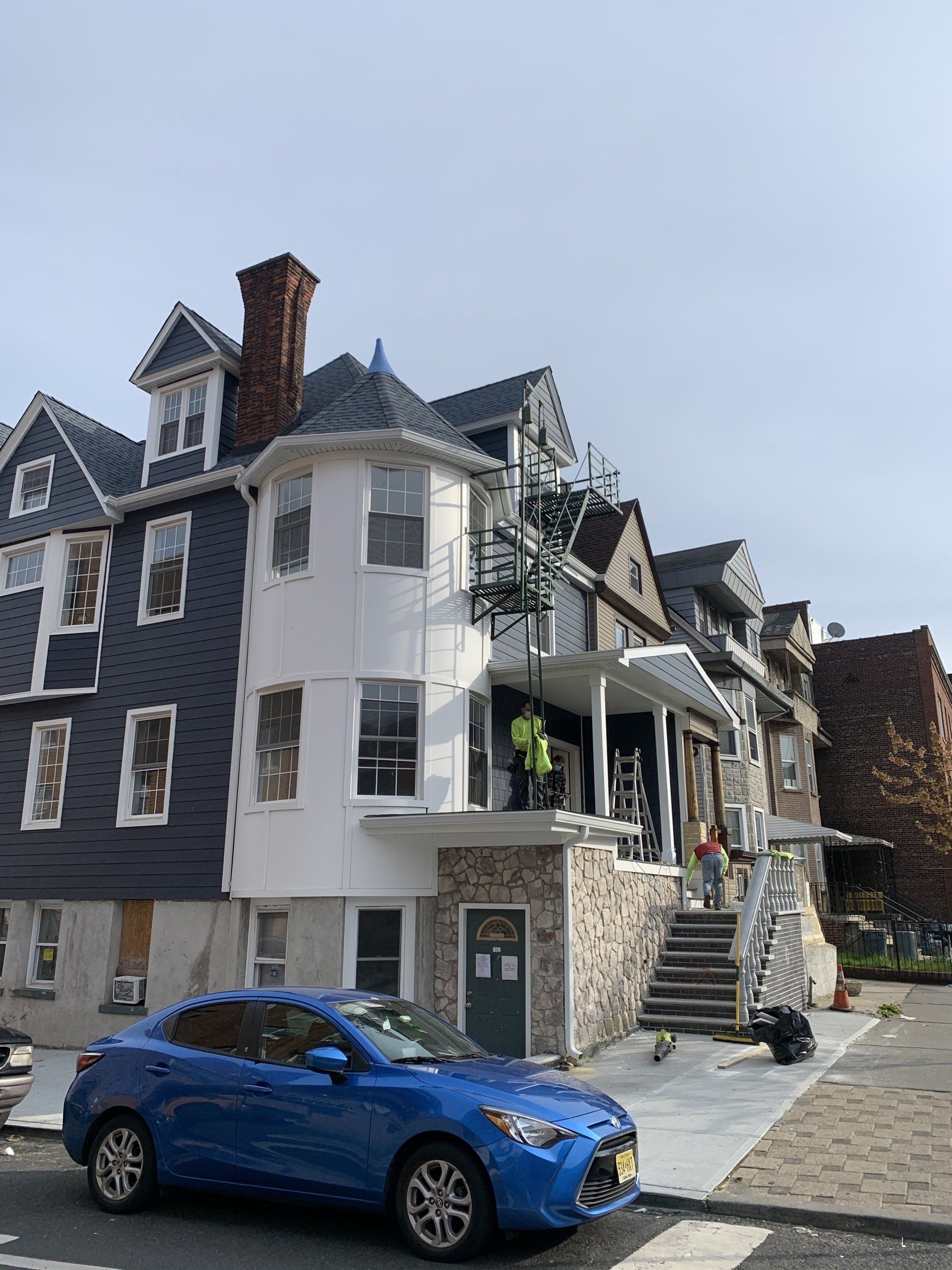 Blue car parked beside a damaged multi-story house with exposed upper floors and a boarded-up entrance