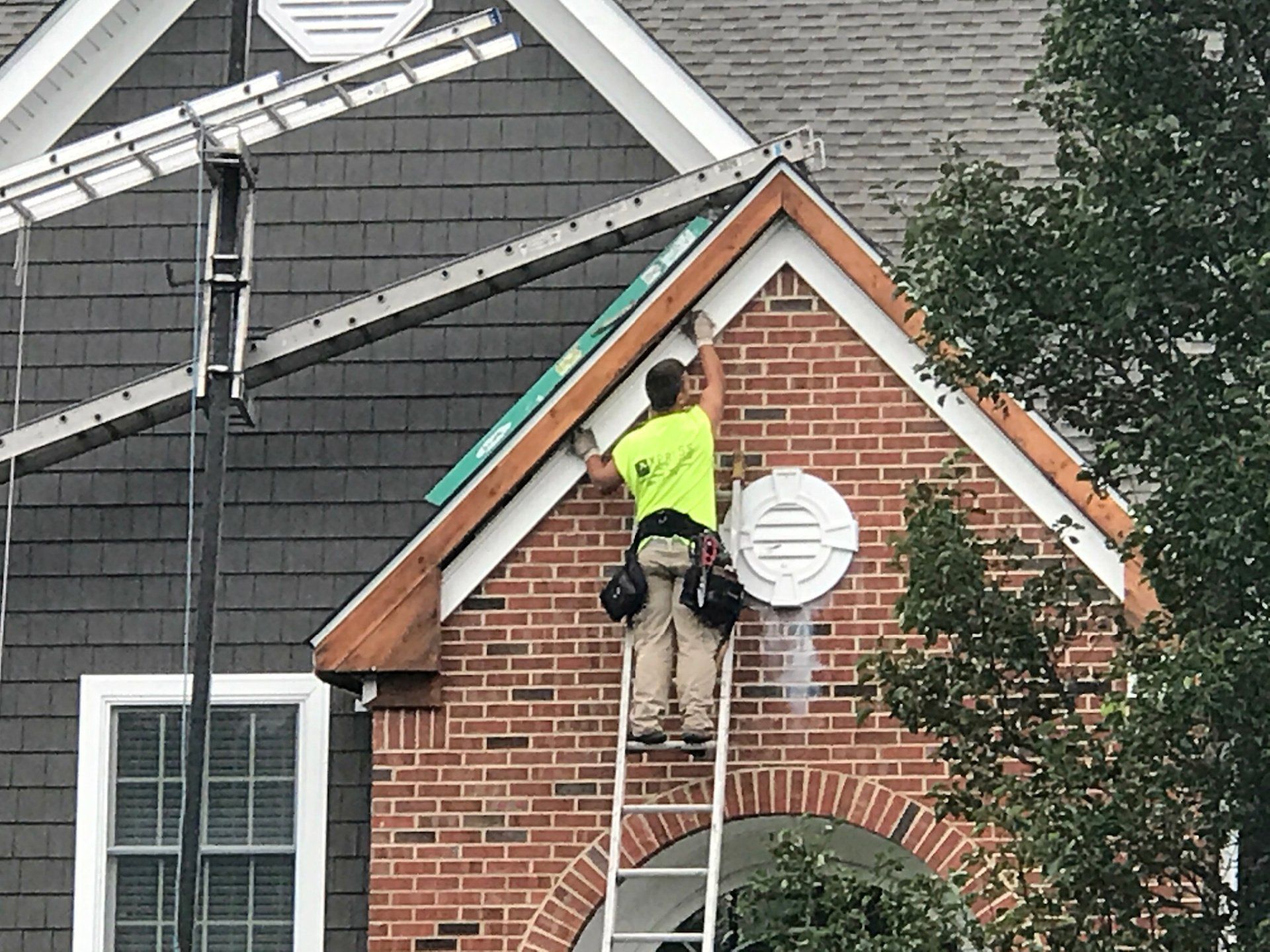 Worker on ladder repairing roof trim on a brick house with white siding and gray shingles