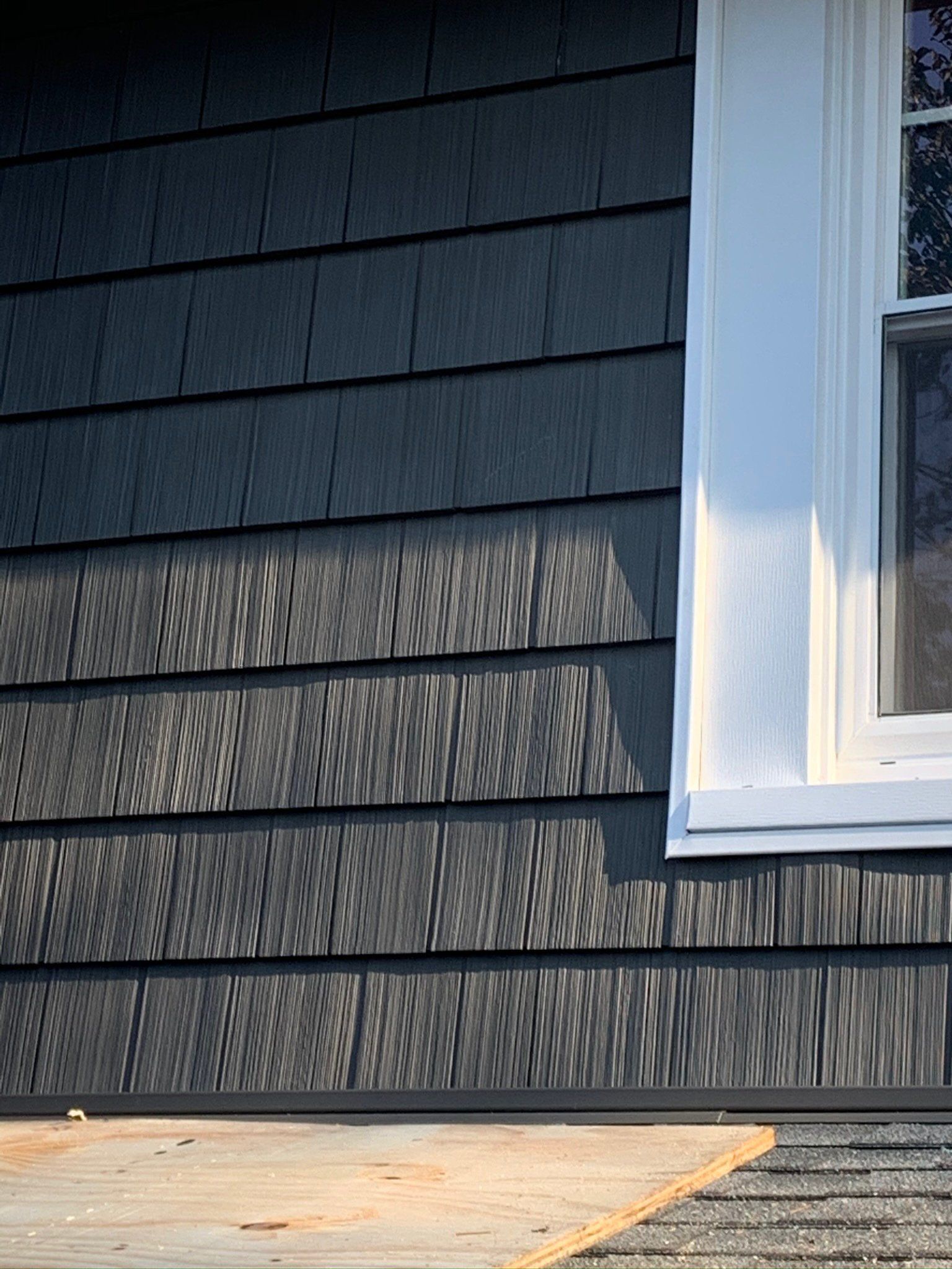 Dark gray house siding with a white window above a sunlit wooden deck.