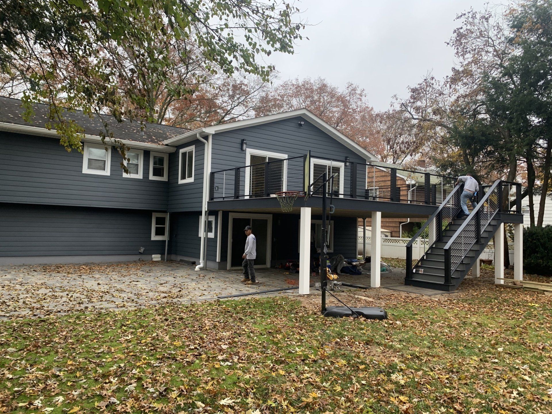 Gray house with raised deck, stairs, and a person standing underneath in a leaf-covered yard