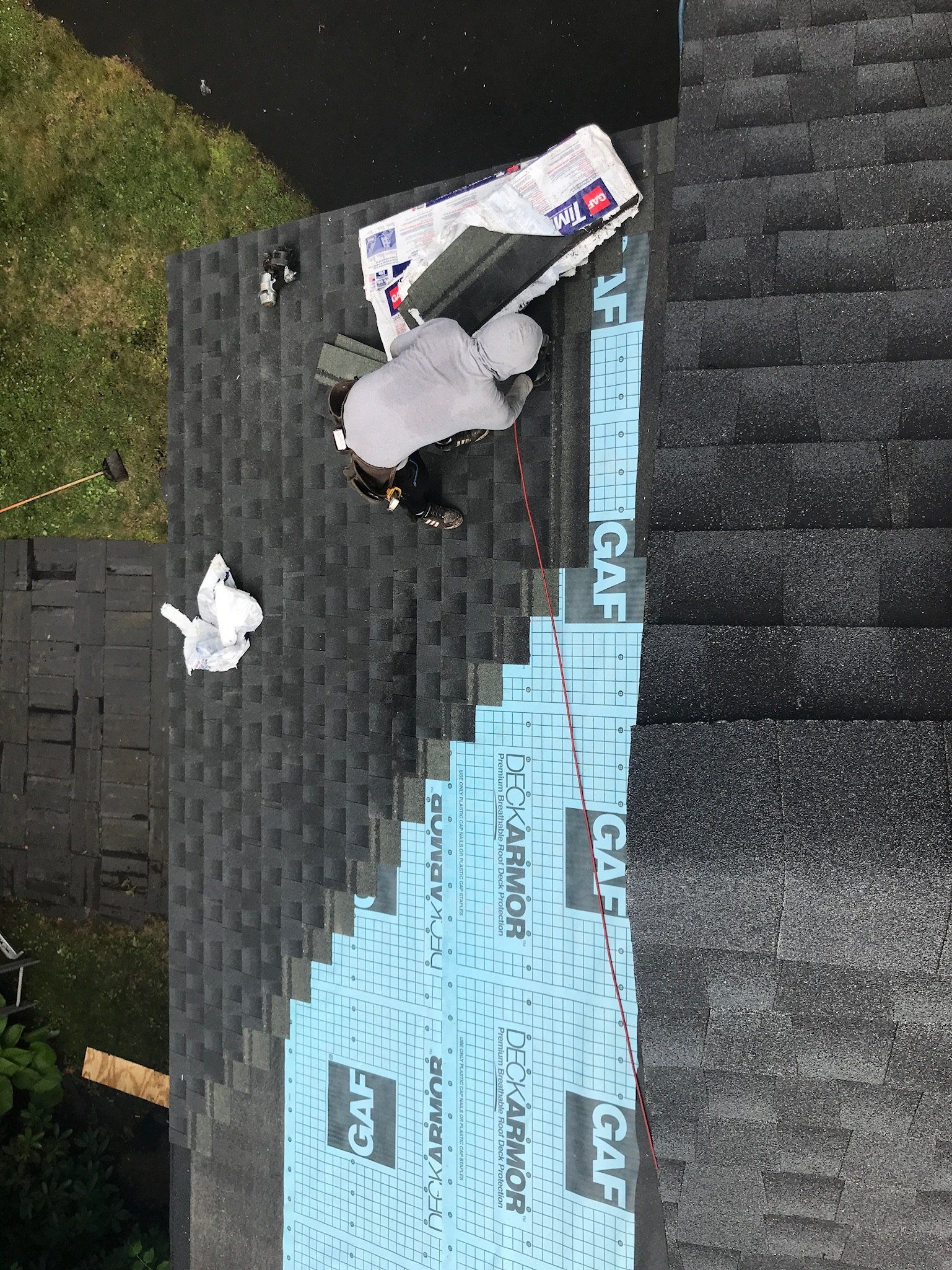 Person working on a roof with blue underlayment and black shingles, near a grassy edge