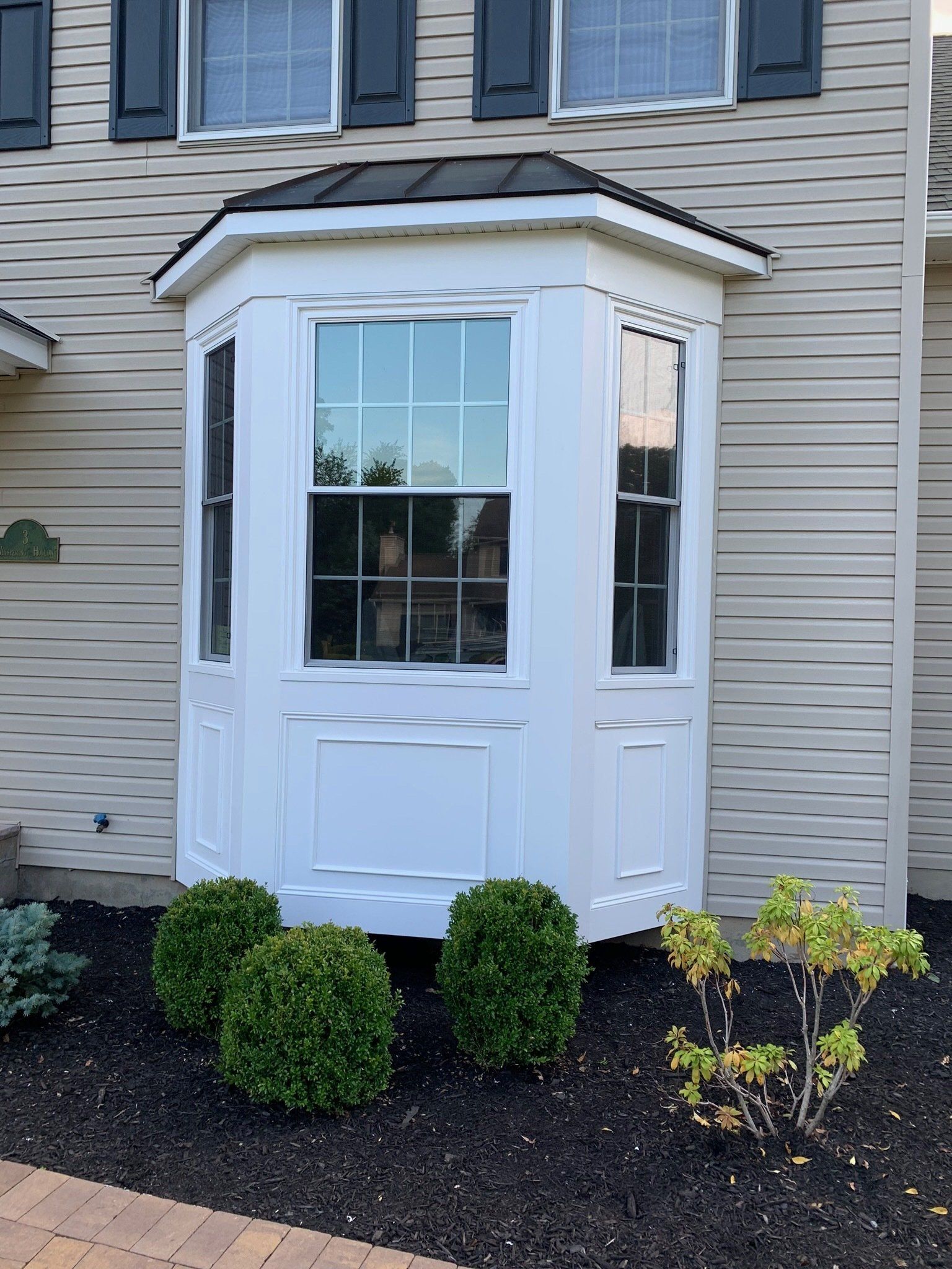 White bay window on beige house exterior with black shutters and shrubs in front