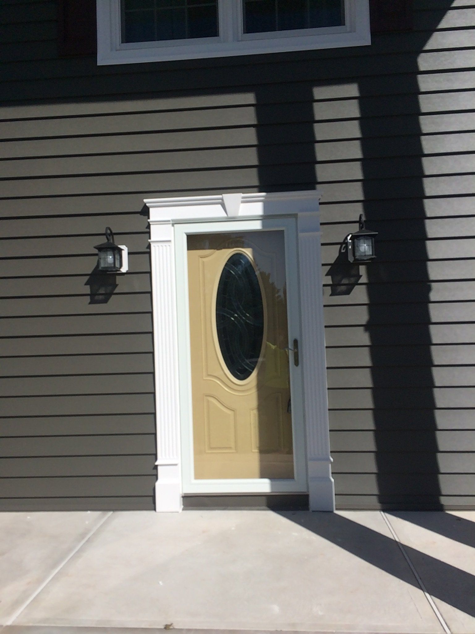Front entrance with yellow door, oval glass, black siding, and two wall lanterns in sunlight