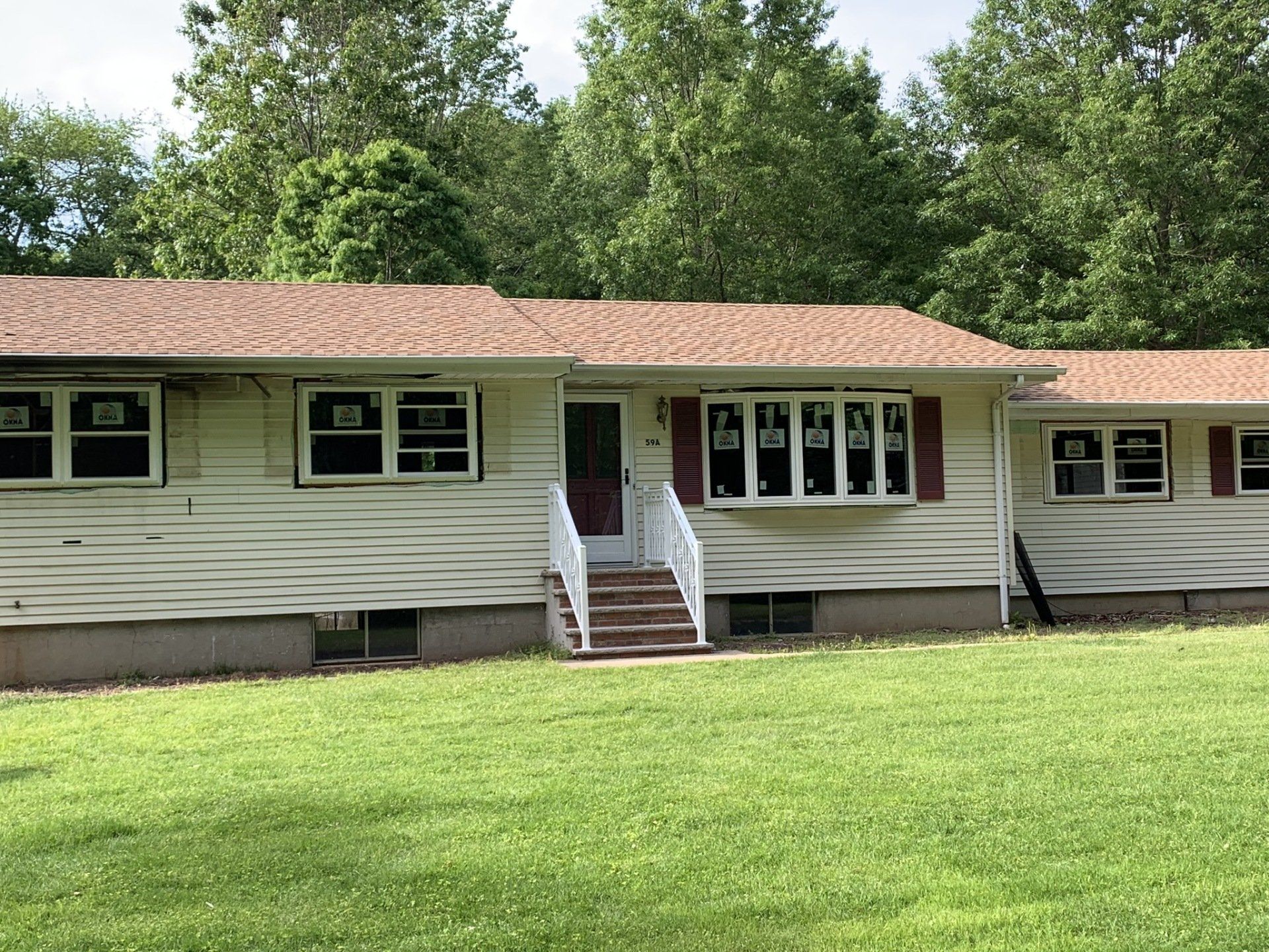 Single-story beige house with red roof, front steps, and green lawn surrounded by trees