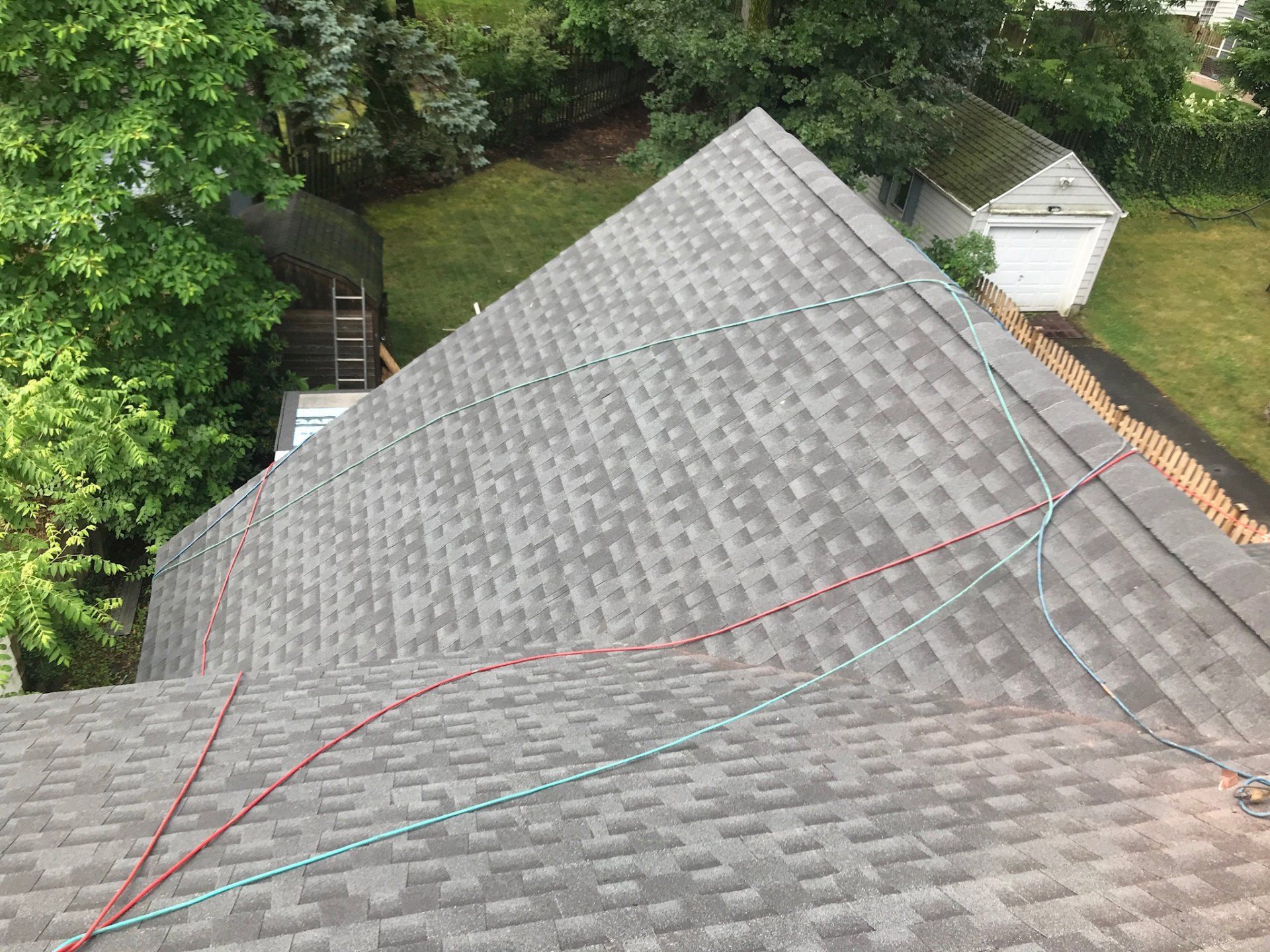 Rooftop with gray shingles viewed from above, with ropes stretching across the roof near trees and a shed.