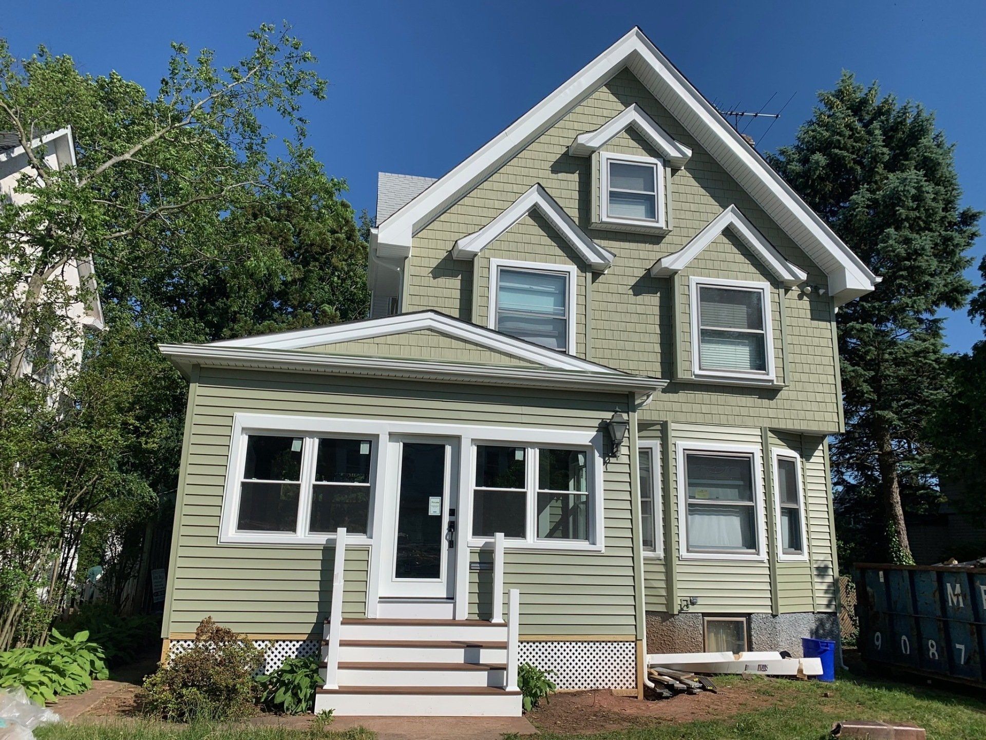 Two-story green house with white trim, front porch, and dormers under a clear blue sky