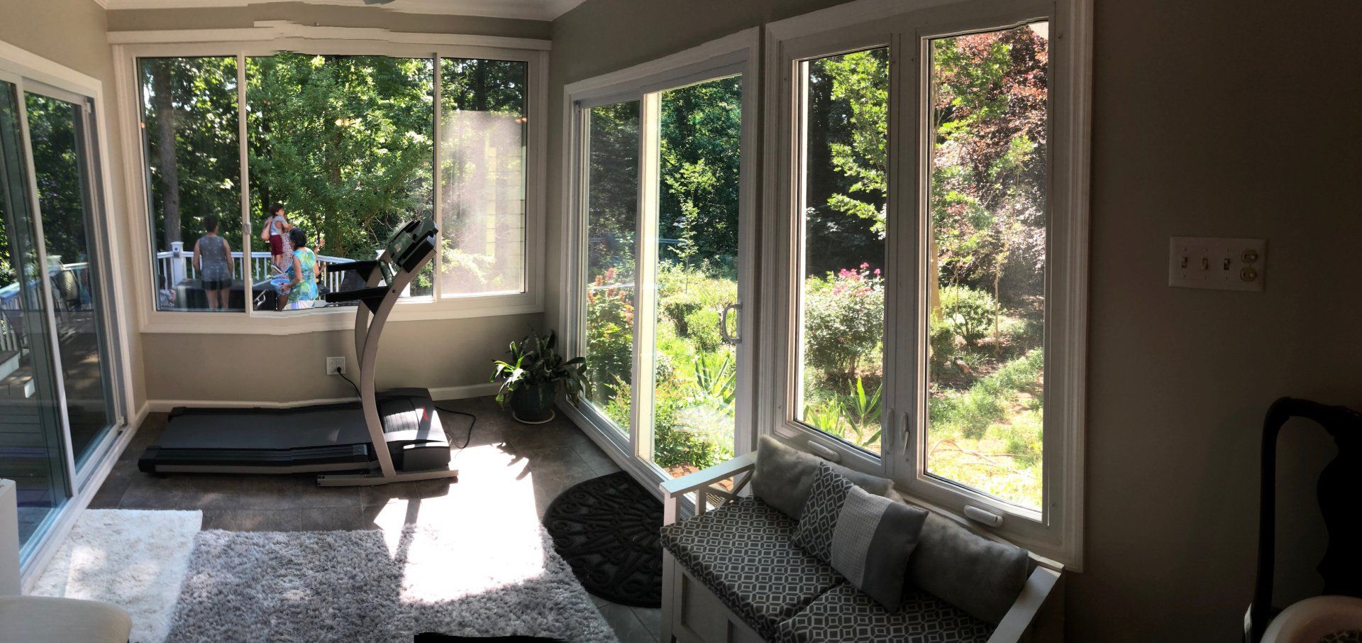 Sunlit living room with large windows, a black treadmill, and a gray couch by the garden view