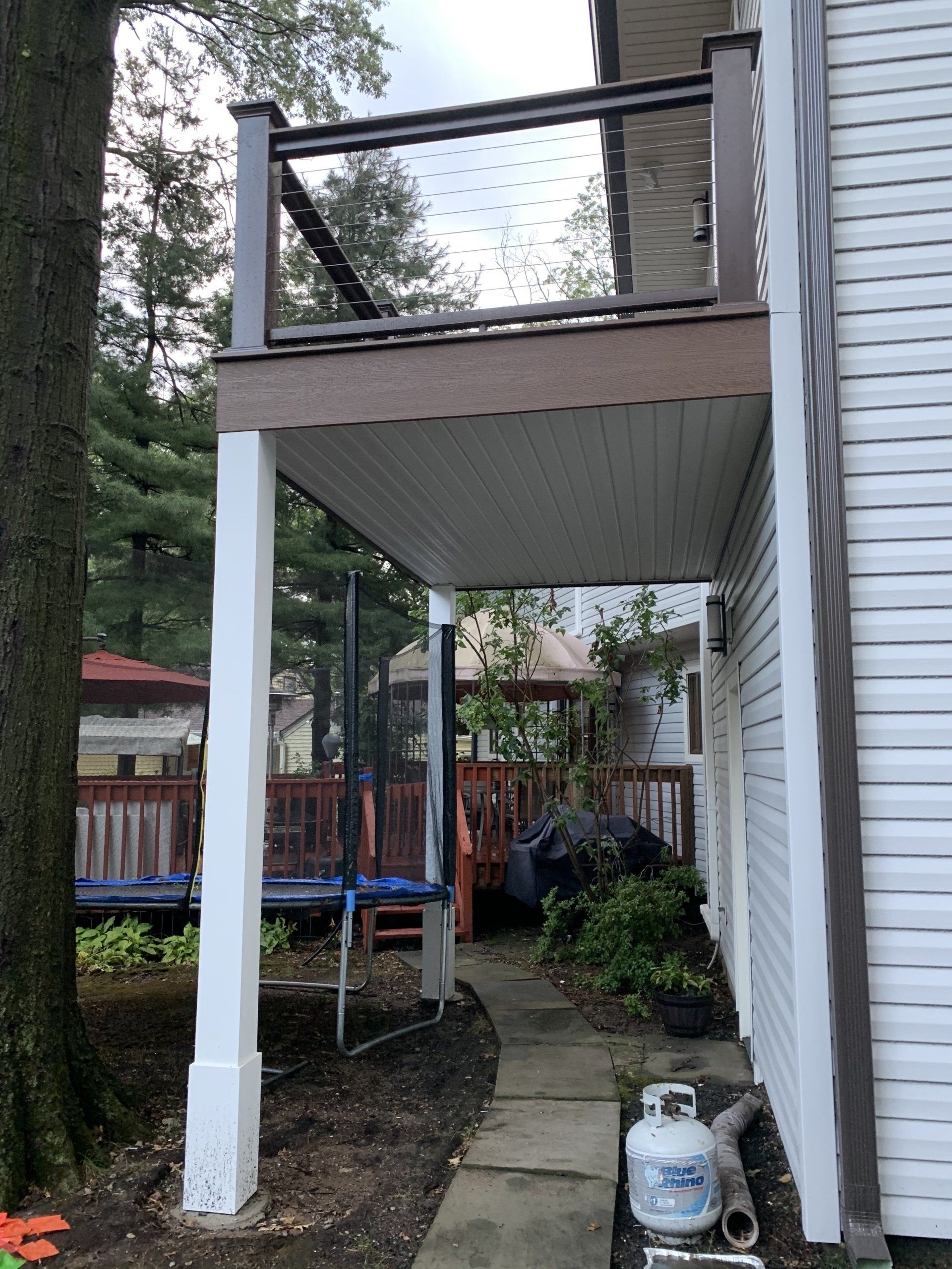 Covered side porch with white posts, brown railing, and a narrow concrete path beside a house.