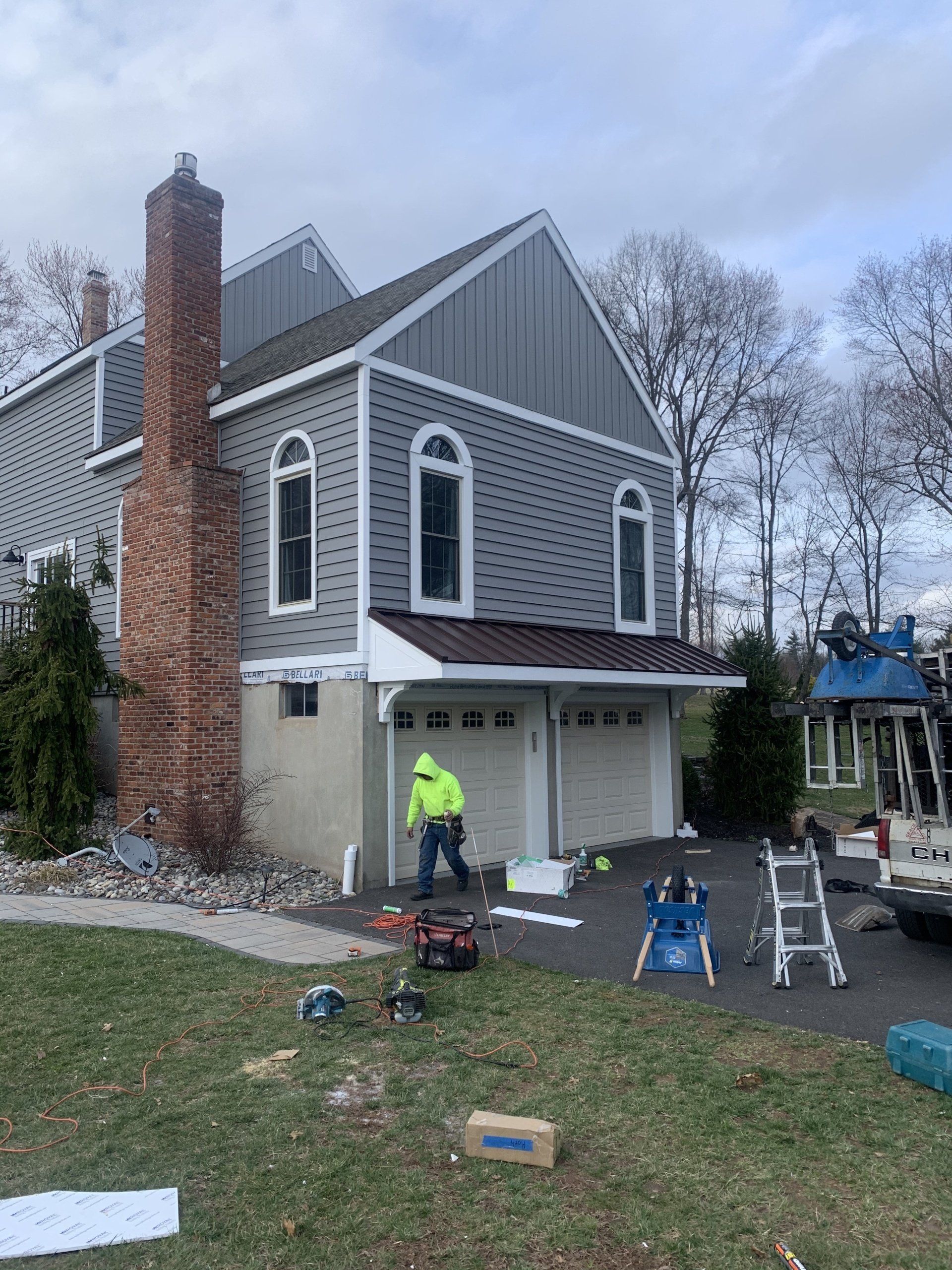 Worker painting the front of a gray house with white trim and a brick chimney.