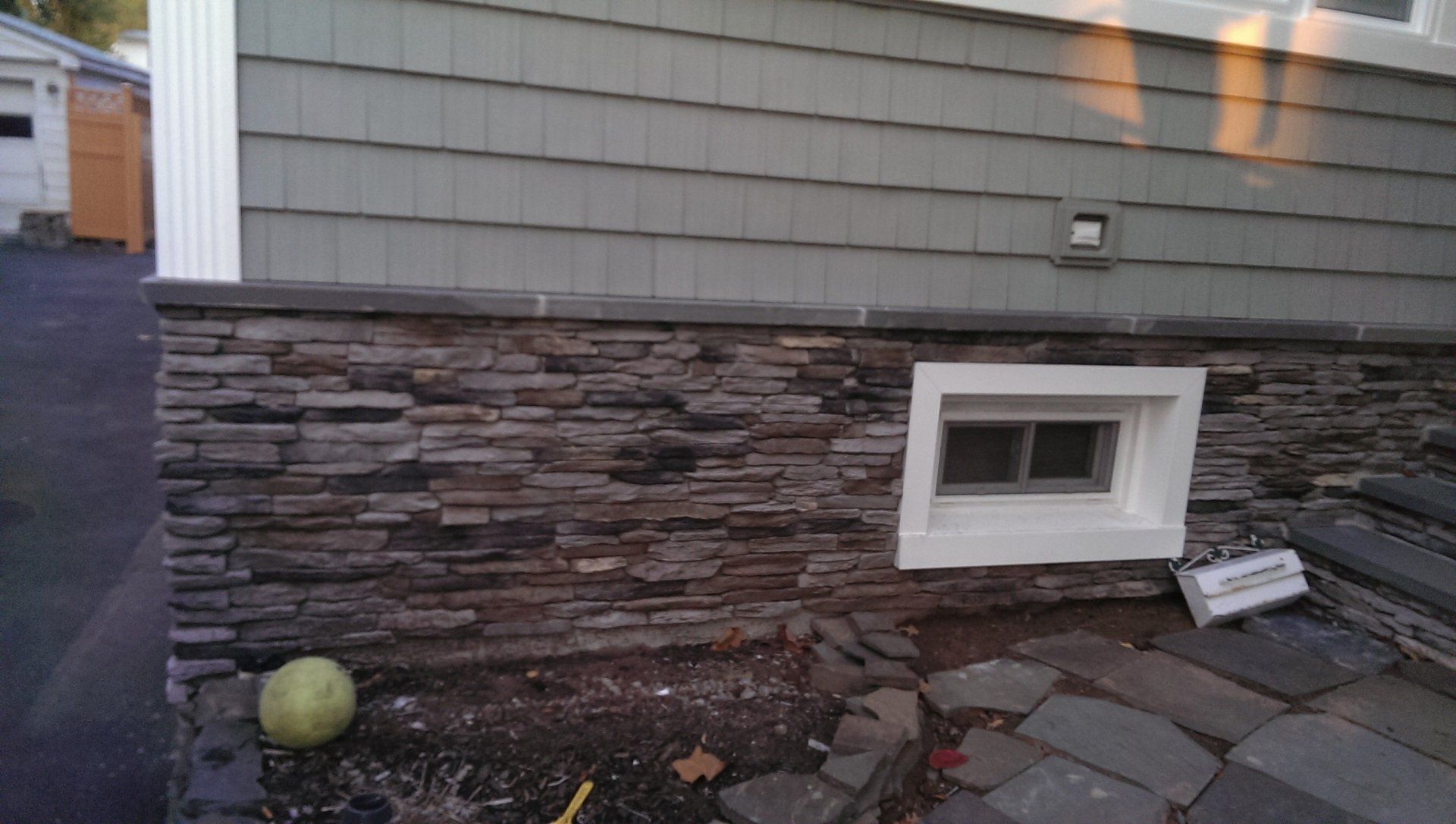 Exterior house wall with stone veneer, small basement window, and potted plant by a stone path