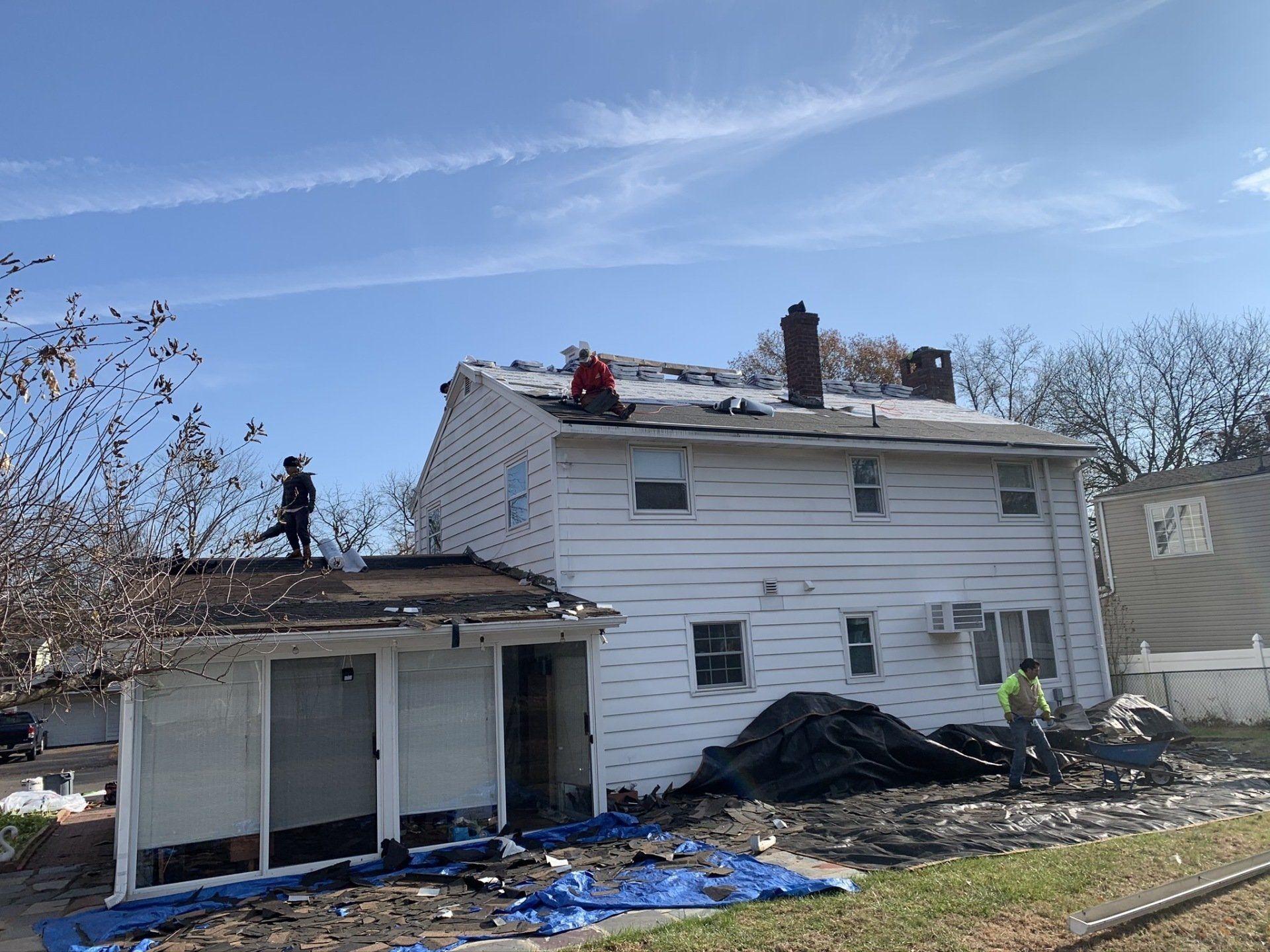 Damaged white house with roof repair workers, tarps, and debris in the yard under a blue sky