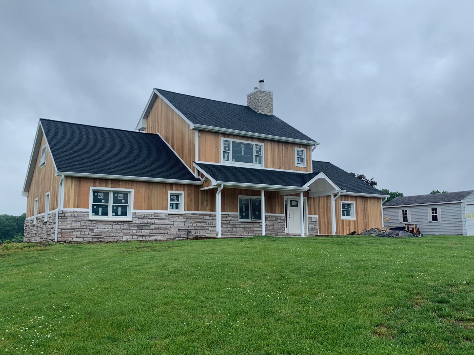 Two-story tan house with stone base and dark roof on a grassy hill under cloudy sky