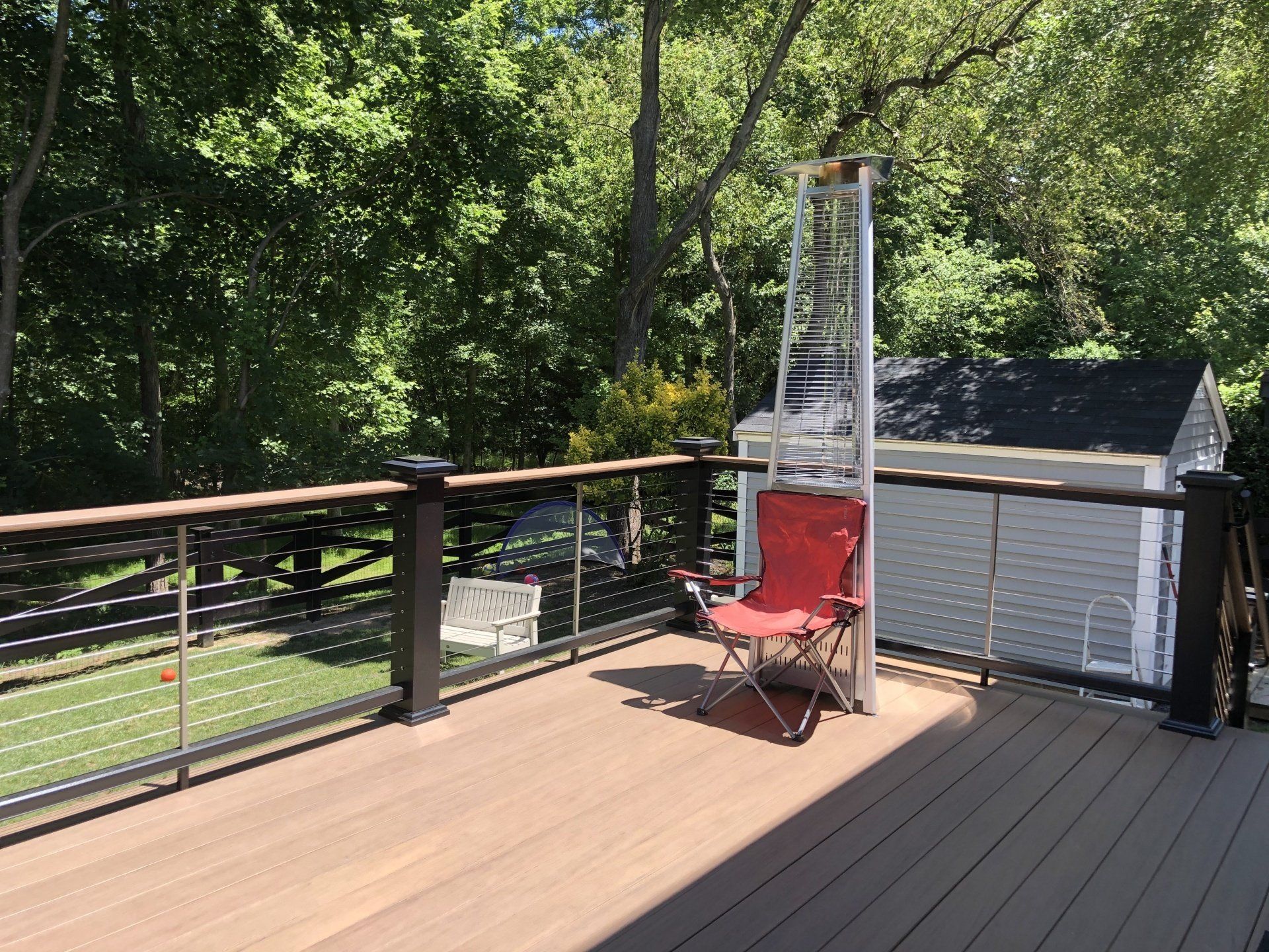 Wooden deck with black railing, red chair, and a tall white metal structure beside a small shed, surrounded by trees