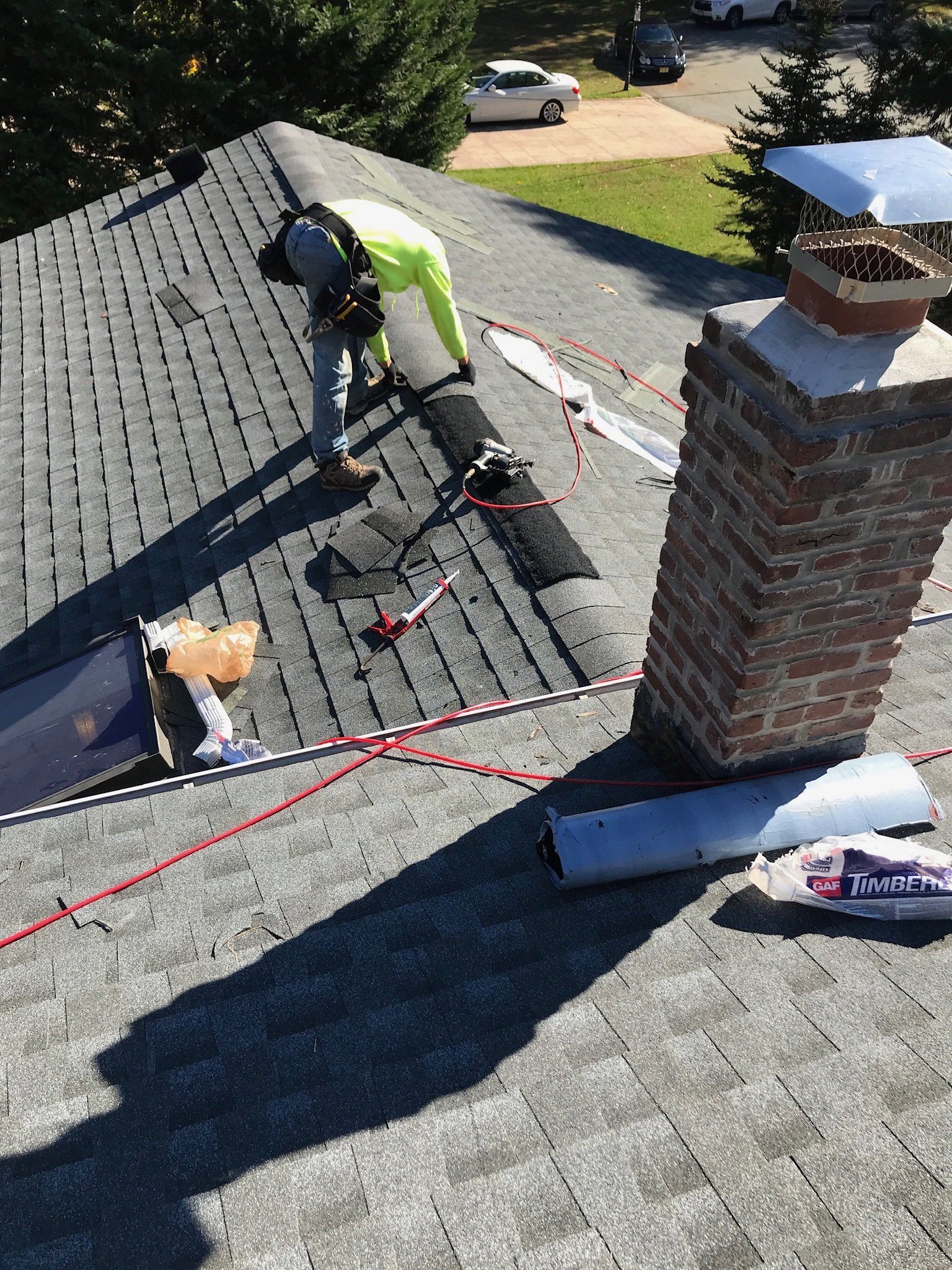 Worker repairing a shingled roof beside a brick chimney, with tools and safety ropes visible.