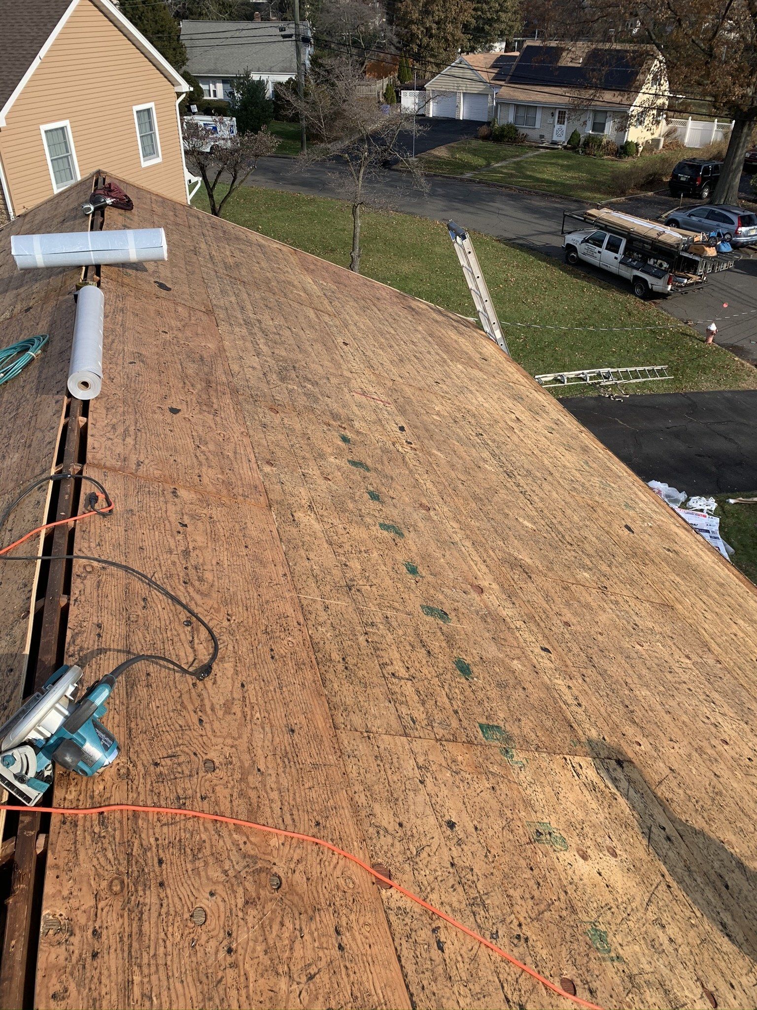 Roofer’s view of a steep shingle roof with tools, orange safety line, and neighboring homes below