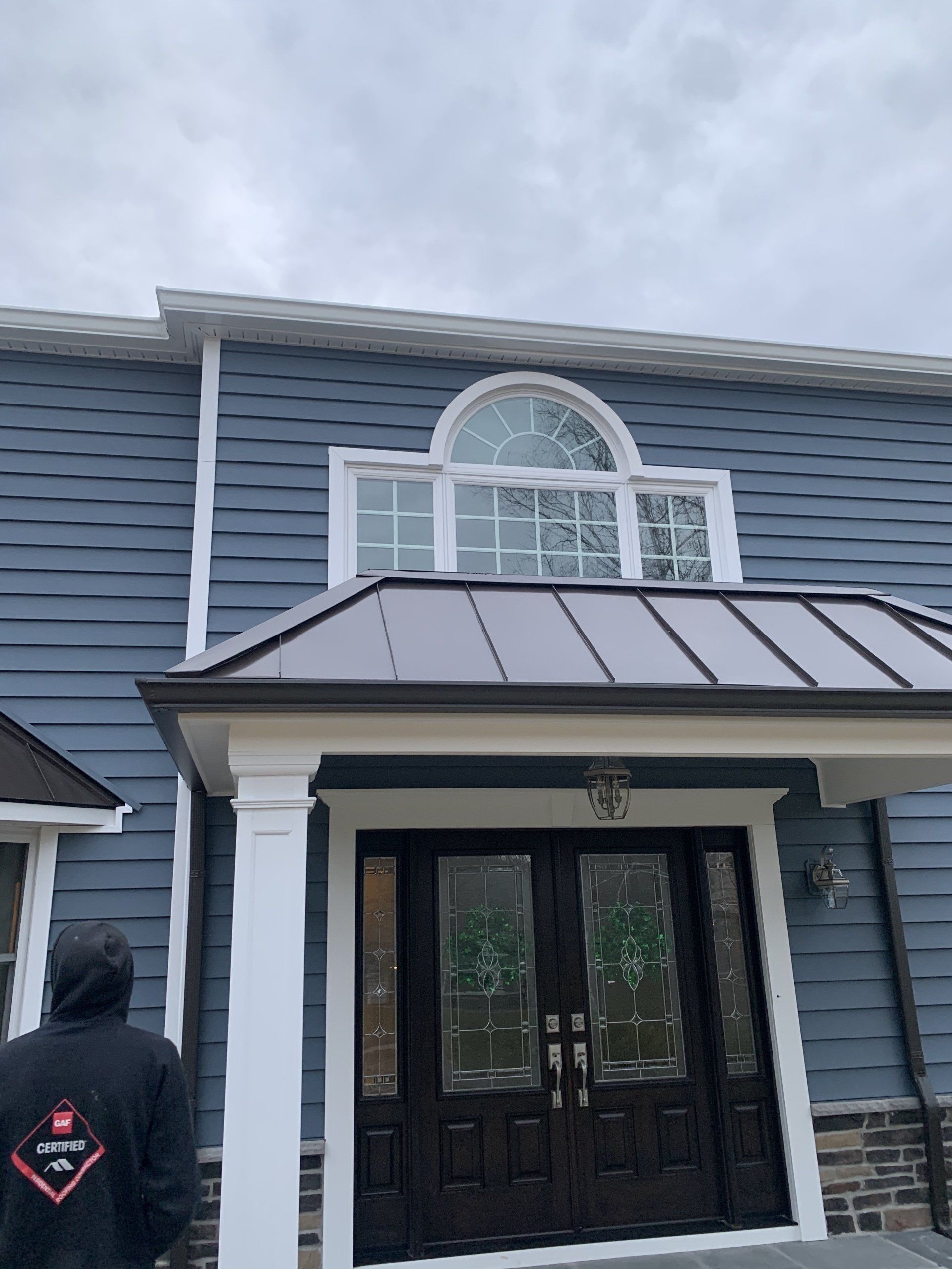 Front entrance of a blue house with white trim, arched window, and black double doors beneath a metal awning.