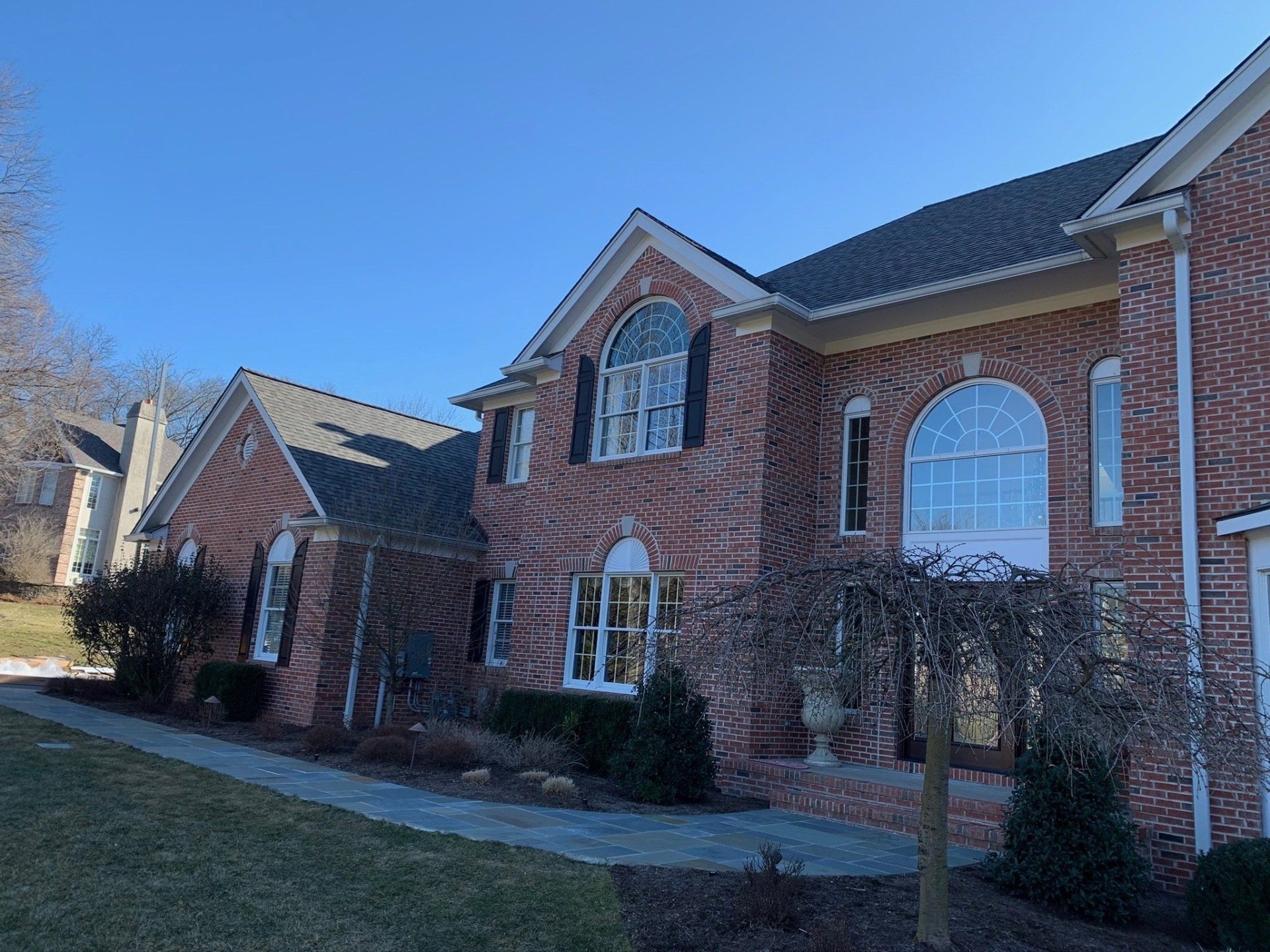 Brick suburban house with arched windows, front walkway, and leafless shrubs on a sunny day