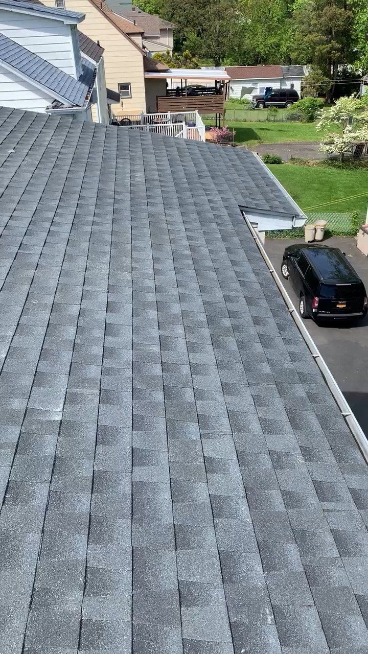 Gray shingle roof beside a driveway, with houses and a green yard in the background.