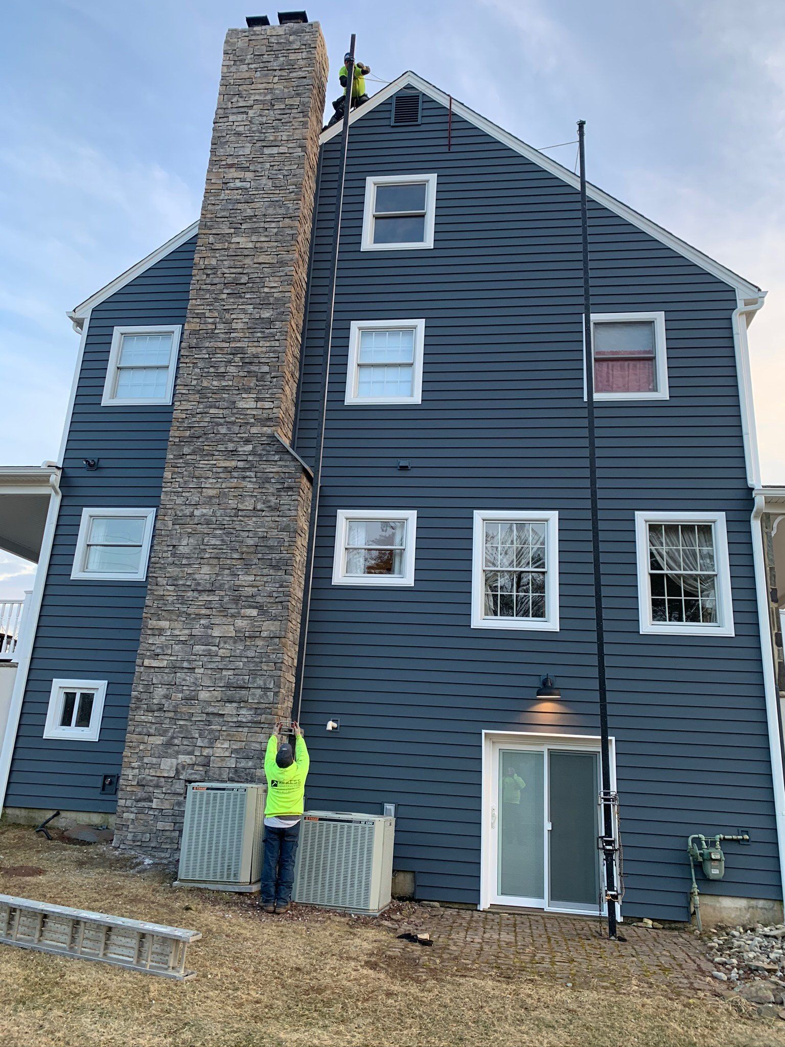 Dark gray multi-story house with a stone chimney, and a worker in a neon jacket standing outside.