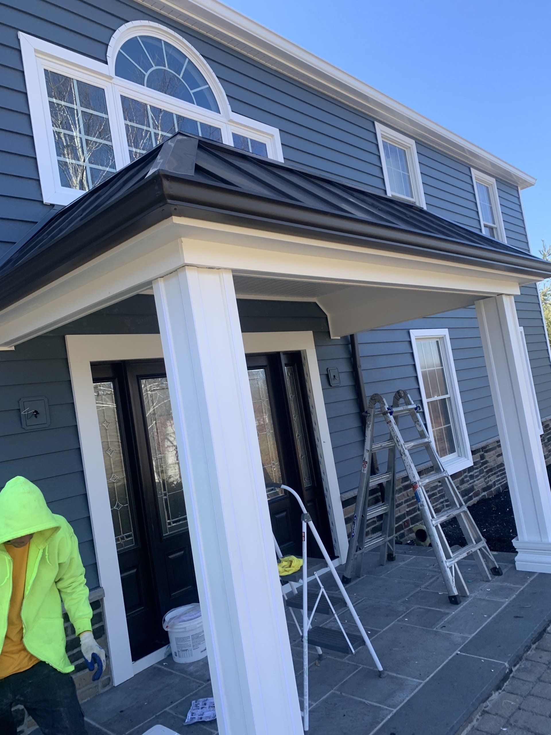 Front porch of a gray house with white columns; workers on ladders painting or repairing siding.