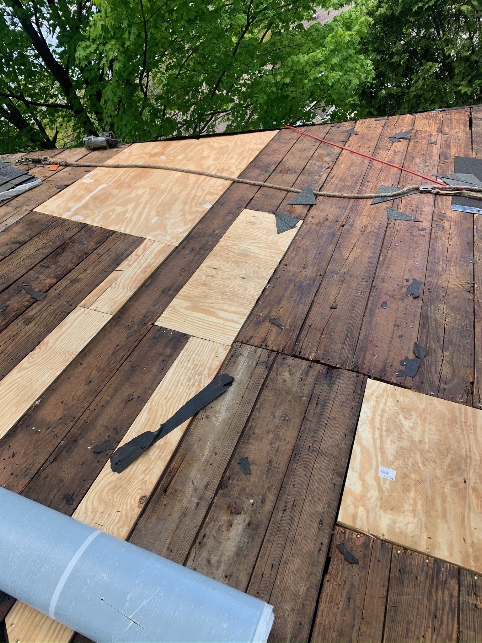 Damaged rooftop with missing shingles and exposed wood near a blue pipe, under green trees