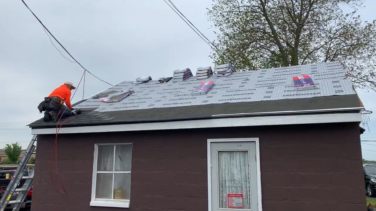 Worker in orange jacket installing shingles on a roof of a small house with a ladder nearby.