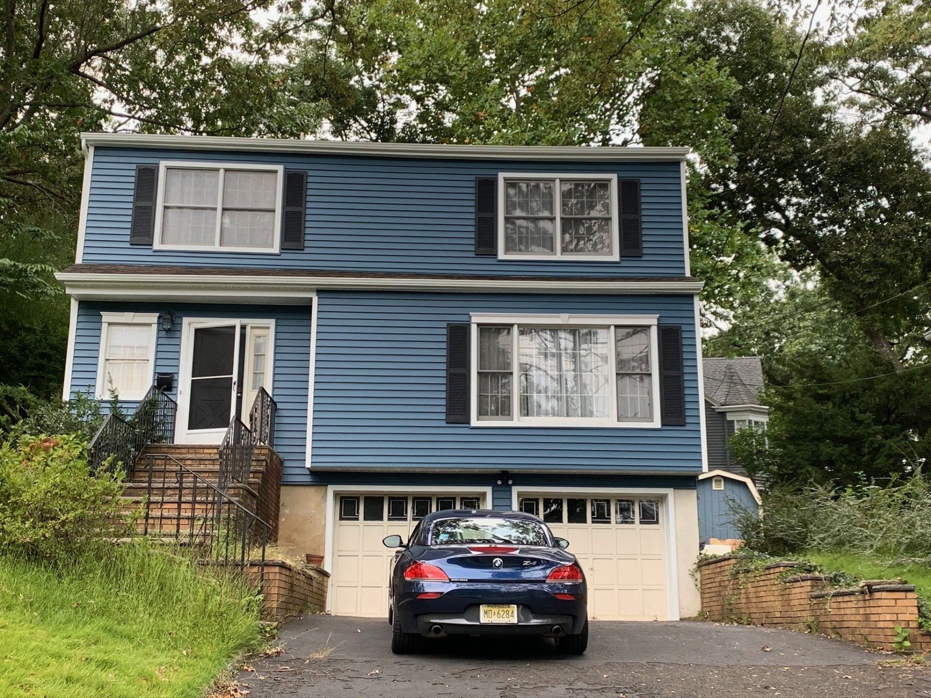 Blue two-story house with white trim, garage, and a black car parked in front, surrounded by trees.