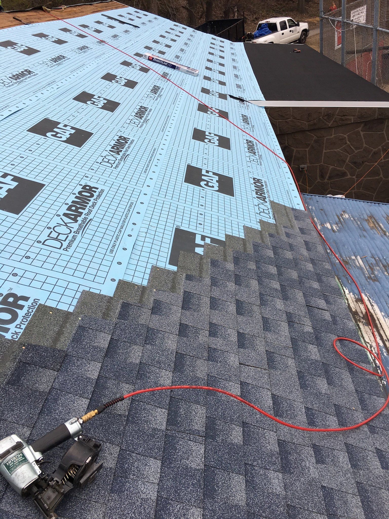 Roof under construction with blue underlayment, gray shingles, and a red hose across the surface
