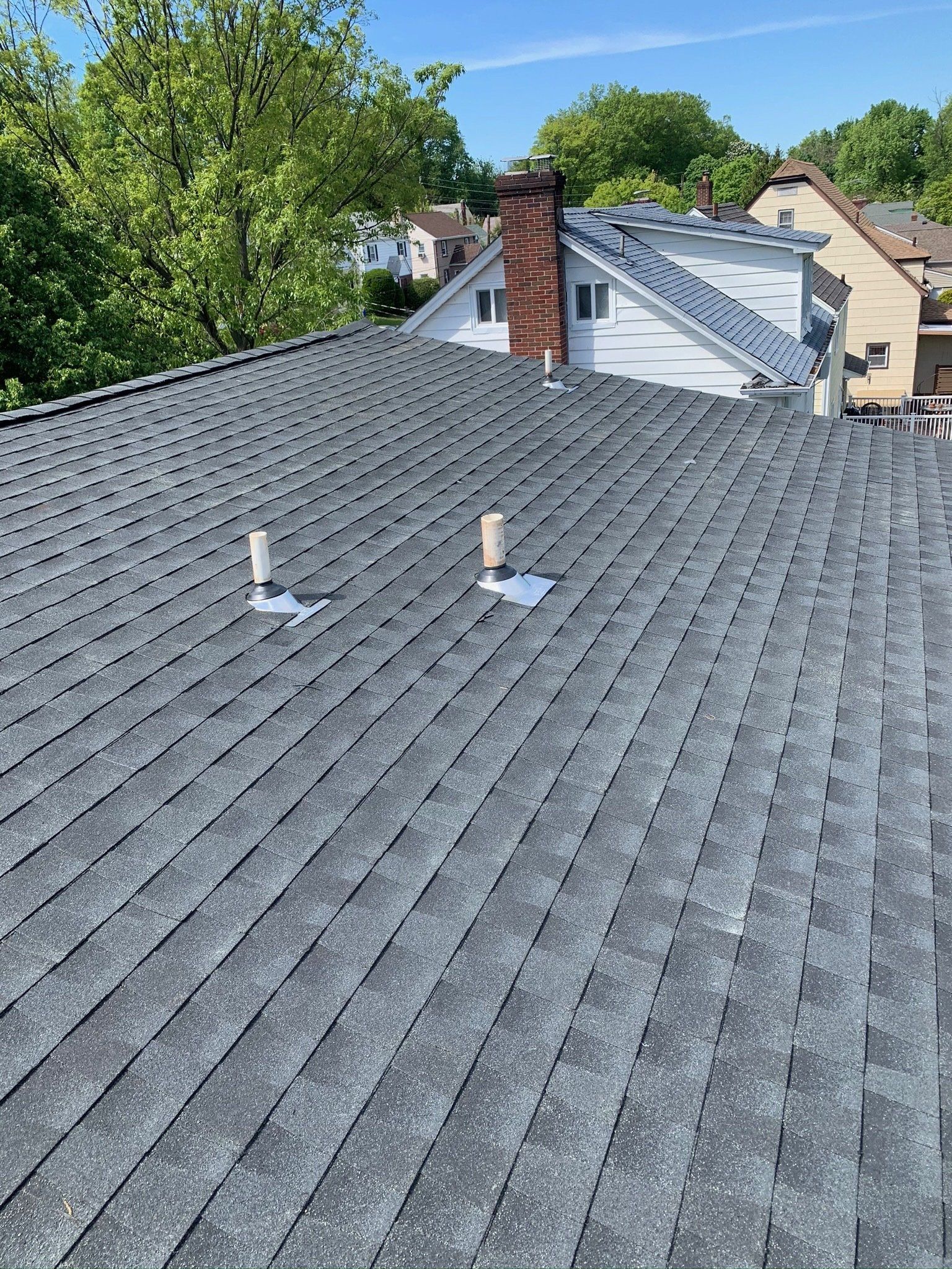 Gray shingled rooftop with two roof vents, trees, and neighboring houses in the background