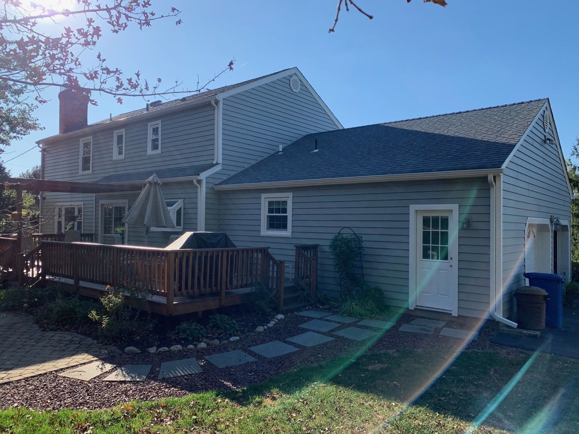 Two-story gray house with a wooden deck, white garage, and driveway in bright sunlight