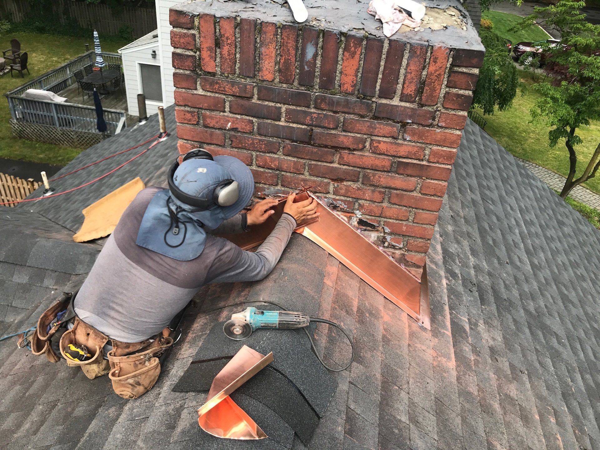 Worker repairing a brick chimney on a shingled roof with tools and safety gear.