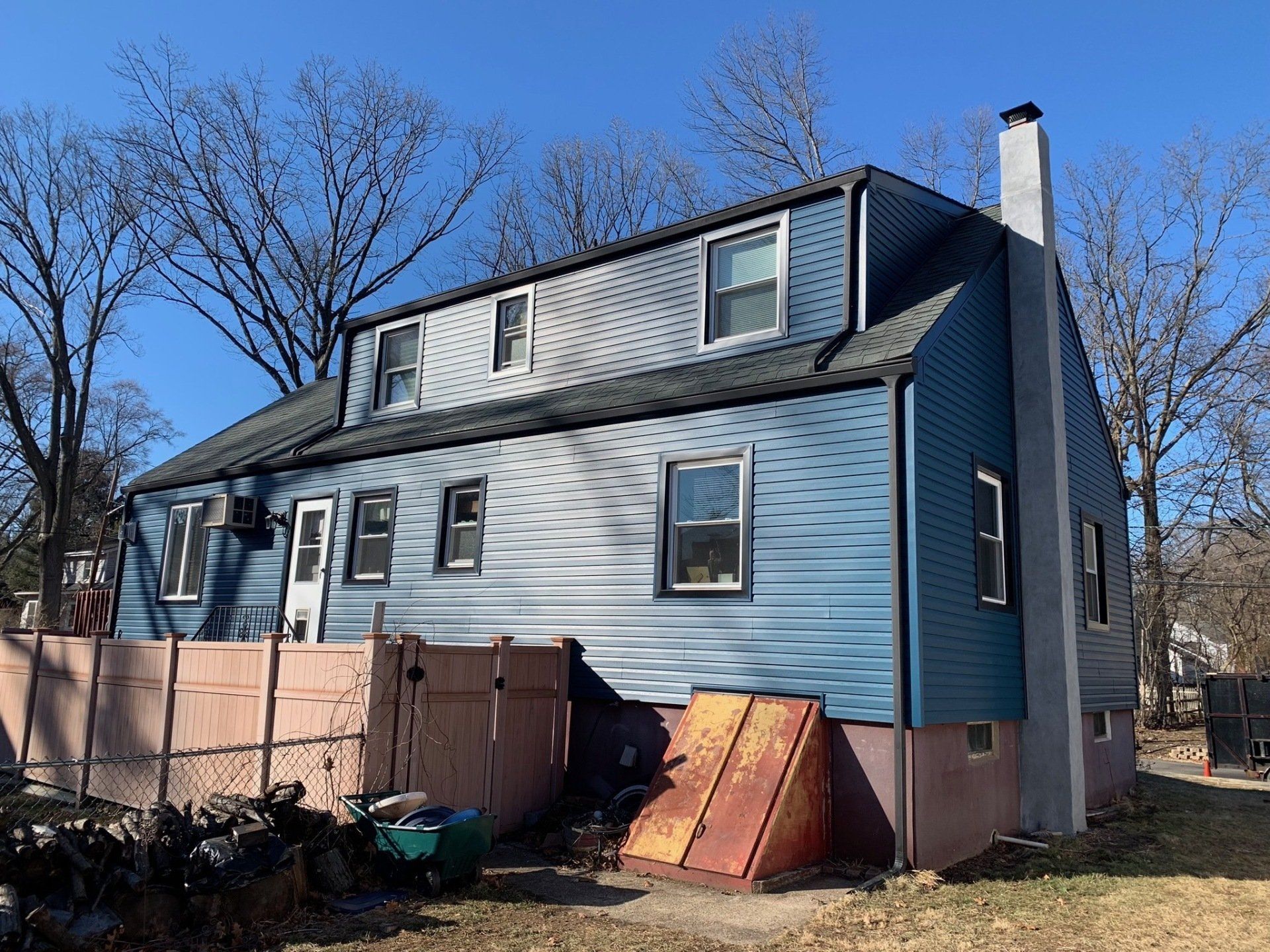 Blue two-story house with dormer windows and a fenced backyard under a clear sky