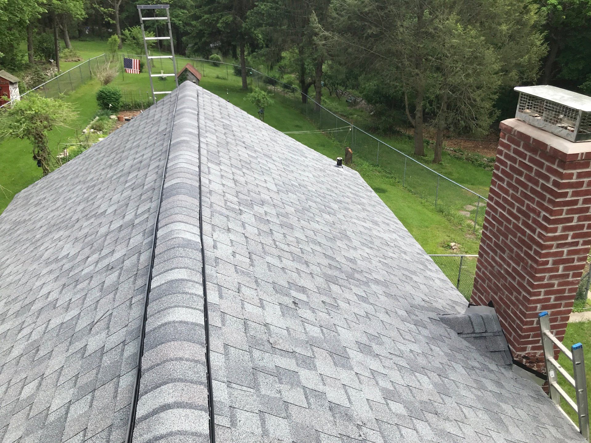 View from a roof ridge with gray shingles, a brick chimney, and green trees and lawn below