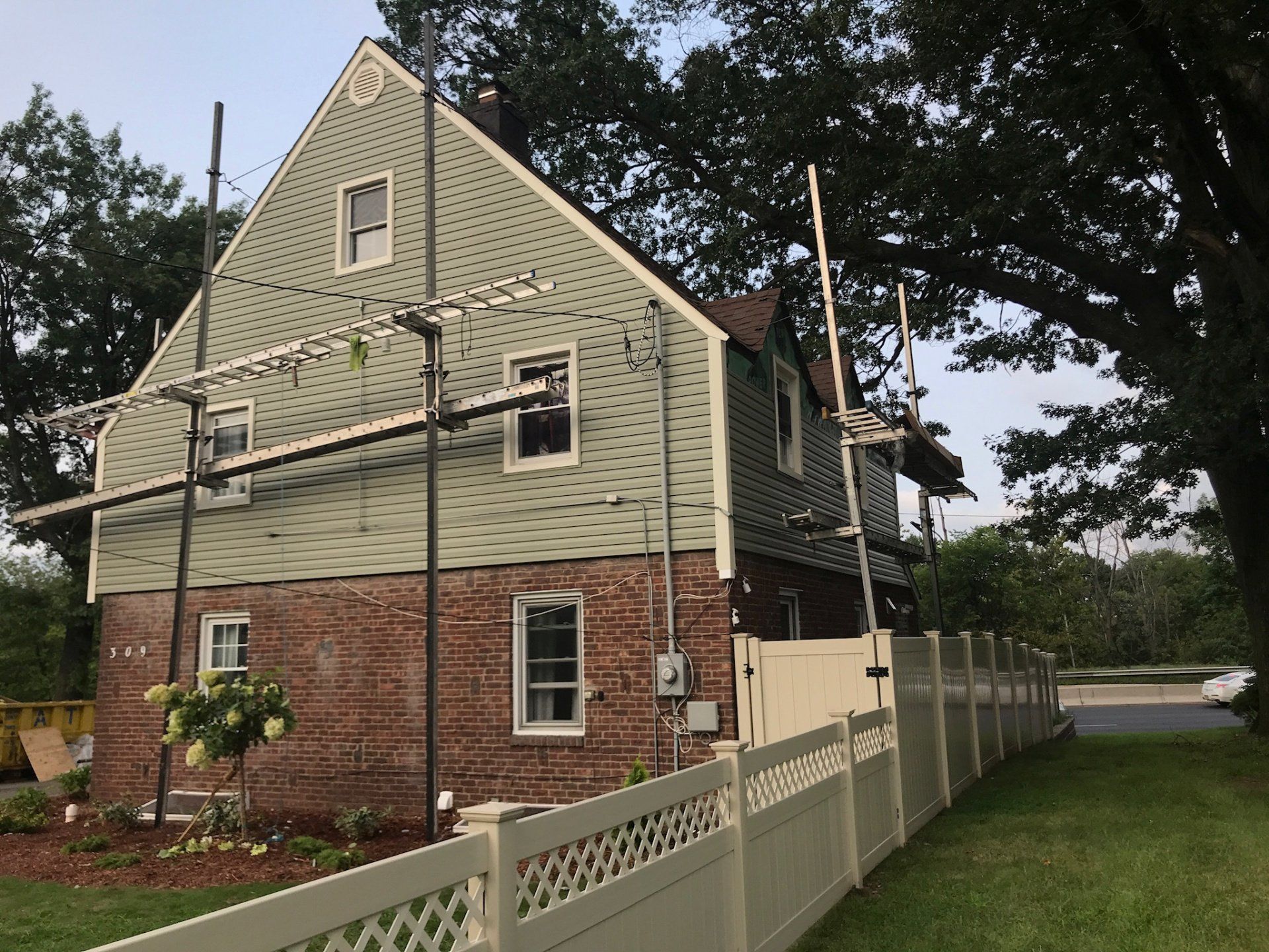 Two-story house with green siding and red brick, surrounded by scaffolding and a white fence