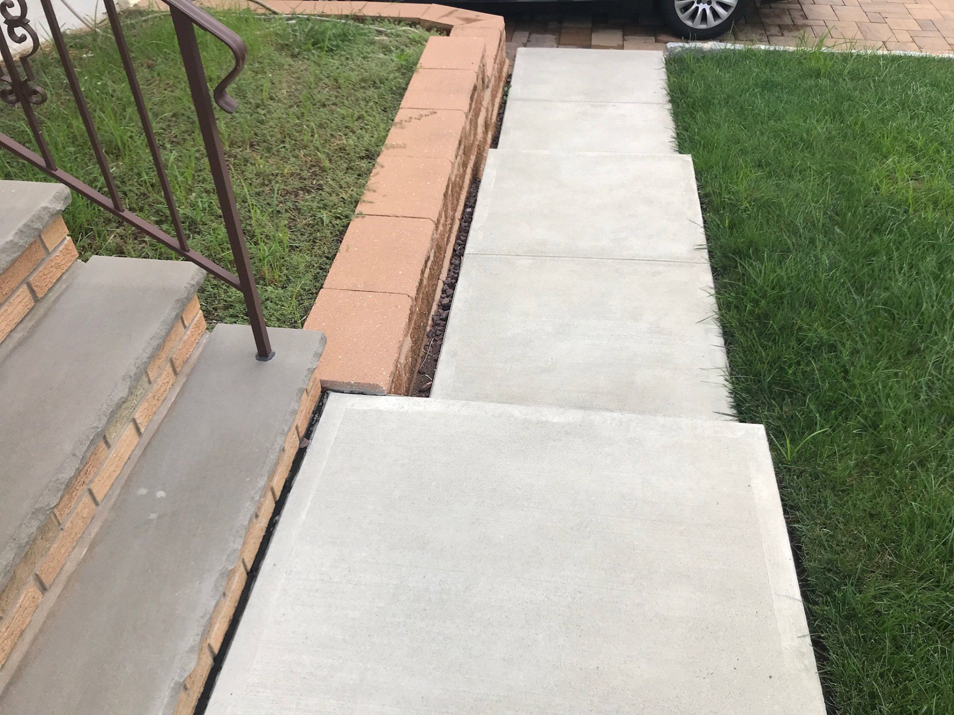 Concrete walkway beside stairs and a grassy lawn, with a brick border and parked car in the distance