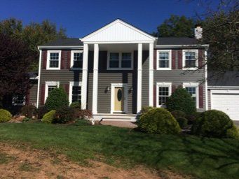 Two-story colonial-style house with white columns, red shutters, and a front lawn under a blue sky