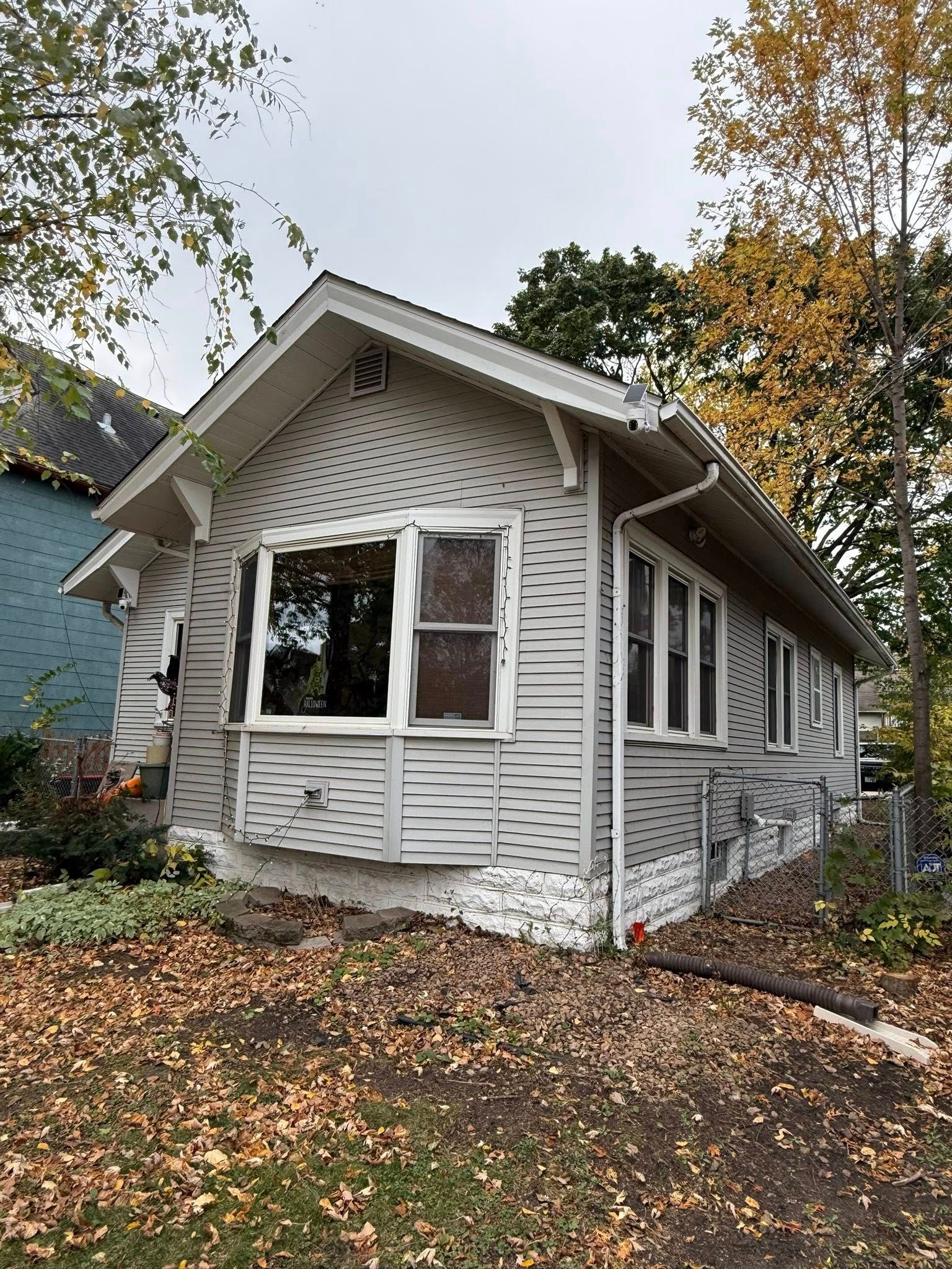 A small, light gray house with a peaked roof and white trim, set among fallen autumn leaves on a cloudy day.