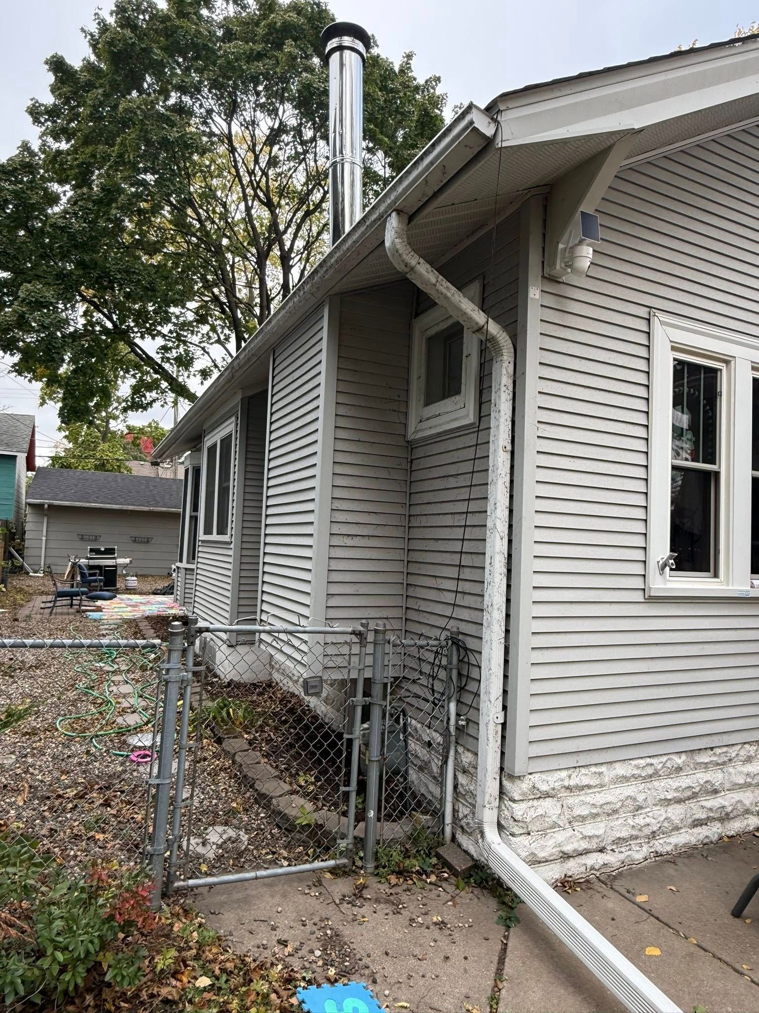 The side exterior of a white, wooden-sided house with a tall metal chimney, a downspout, and a chain-link fence.
