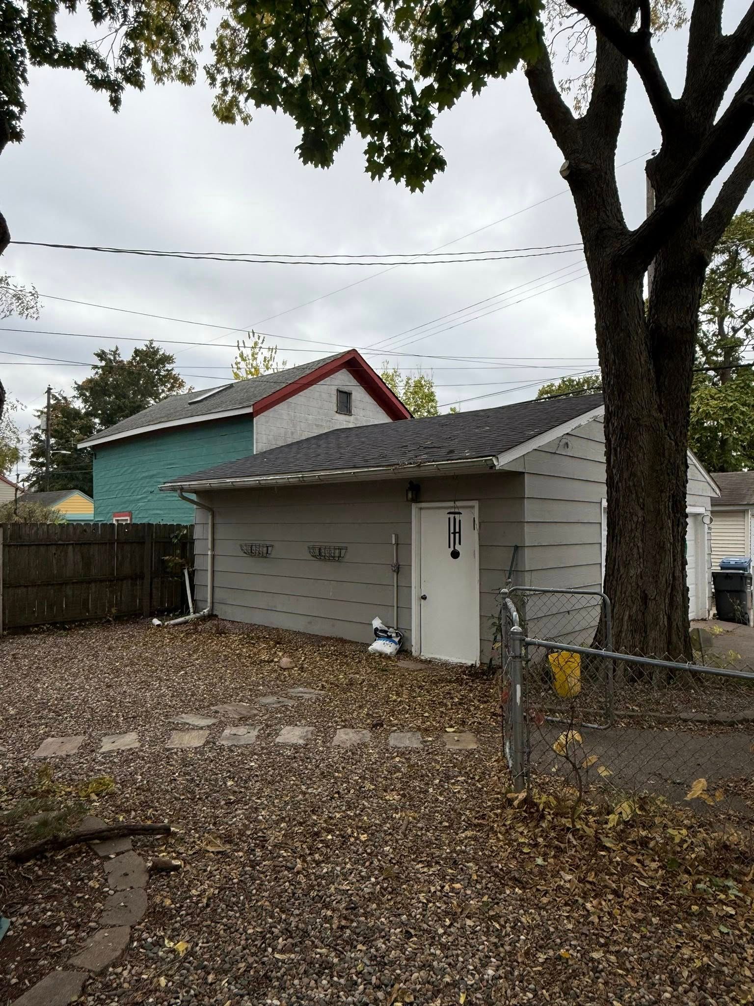 A grey detached garage sits on a gravel lot covered in fallen leaves, beside a turquoise house and a large tree.