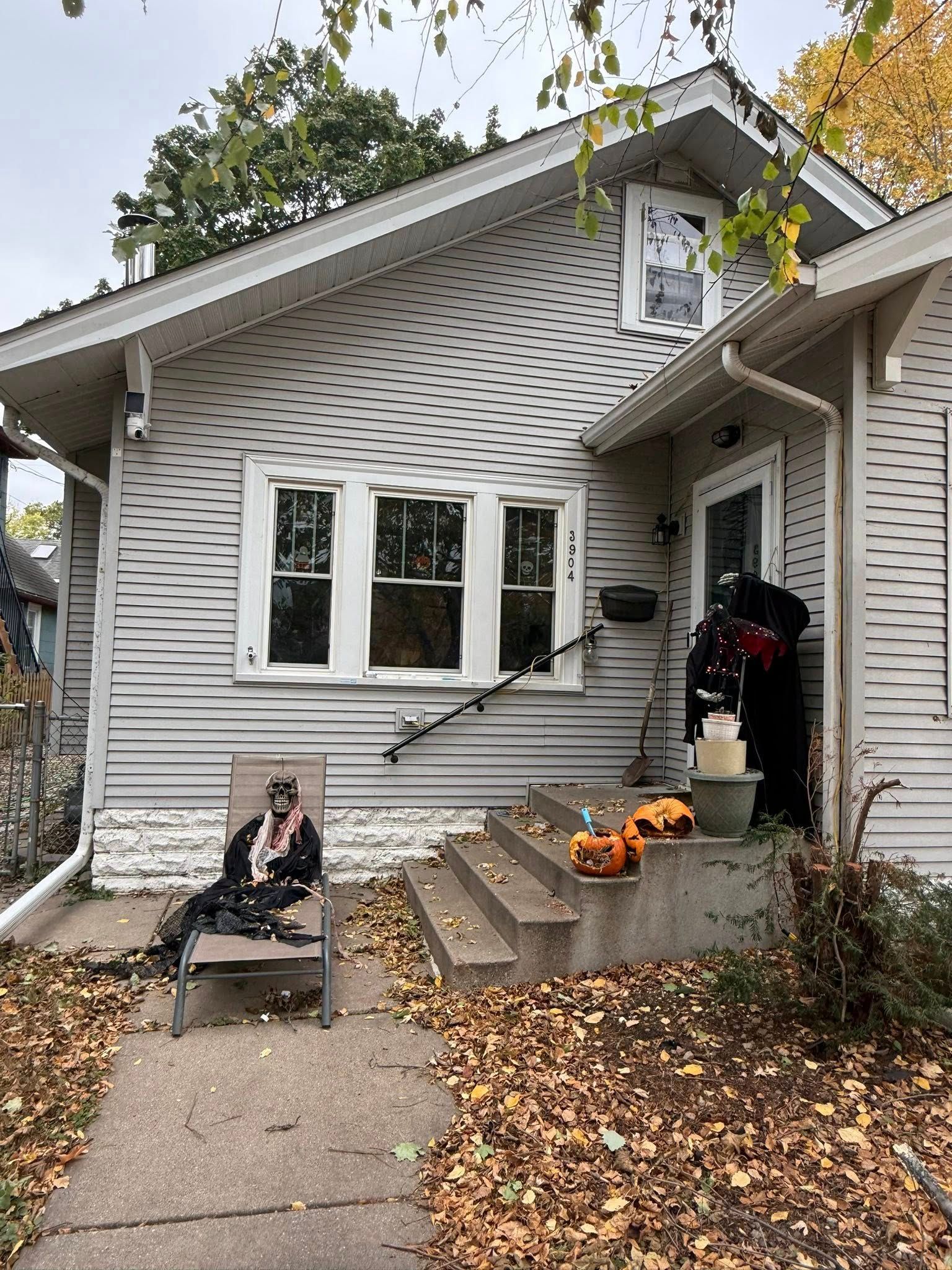 A small house decorated for Halloween with a figure in a lounge chair, pumpkins on the steps, and autumn leaves.