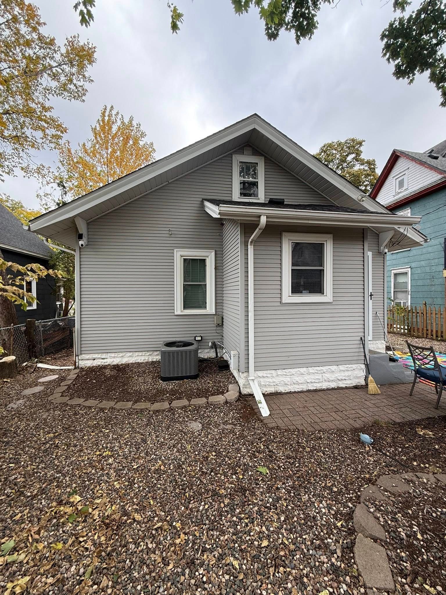 The back of a gray, single-story house with white trim, a small attic window, and an outdoor HVAC unit on a leafy yard.