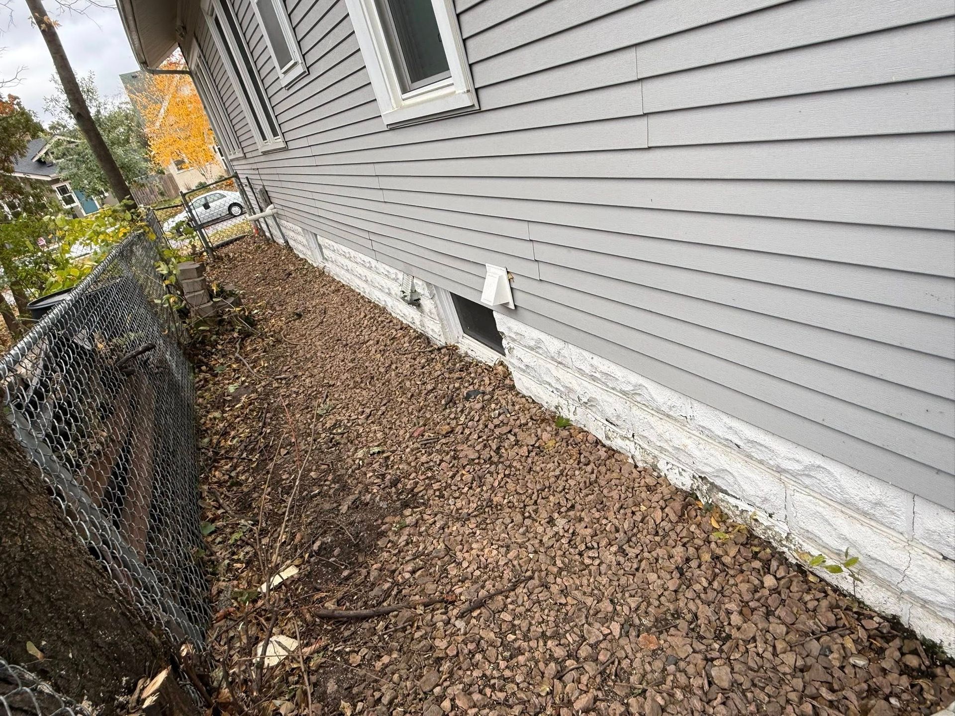 A side view of a house with light gray siding and a stone foundation, next to a gravel path and a chain-link fence.
