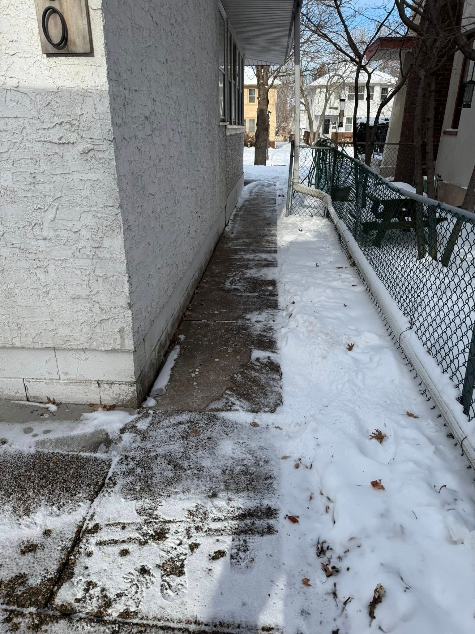 A narrow concrete walkway between a light-colored stucco building and a chain-link fence, partially covered in snow.